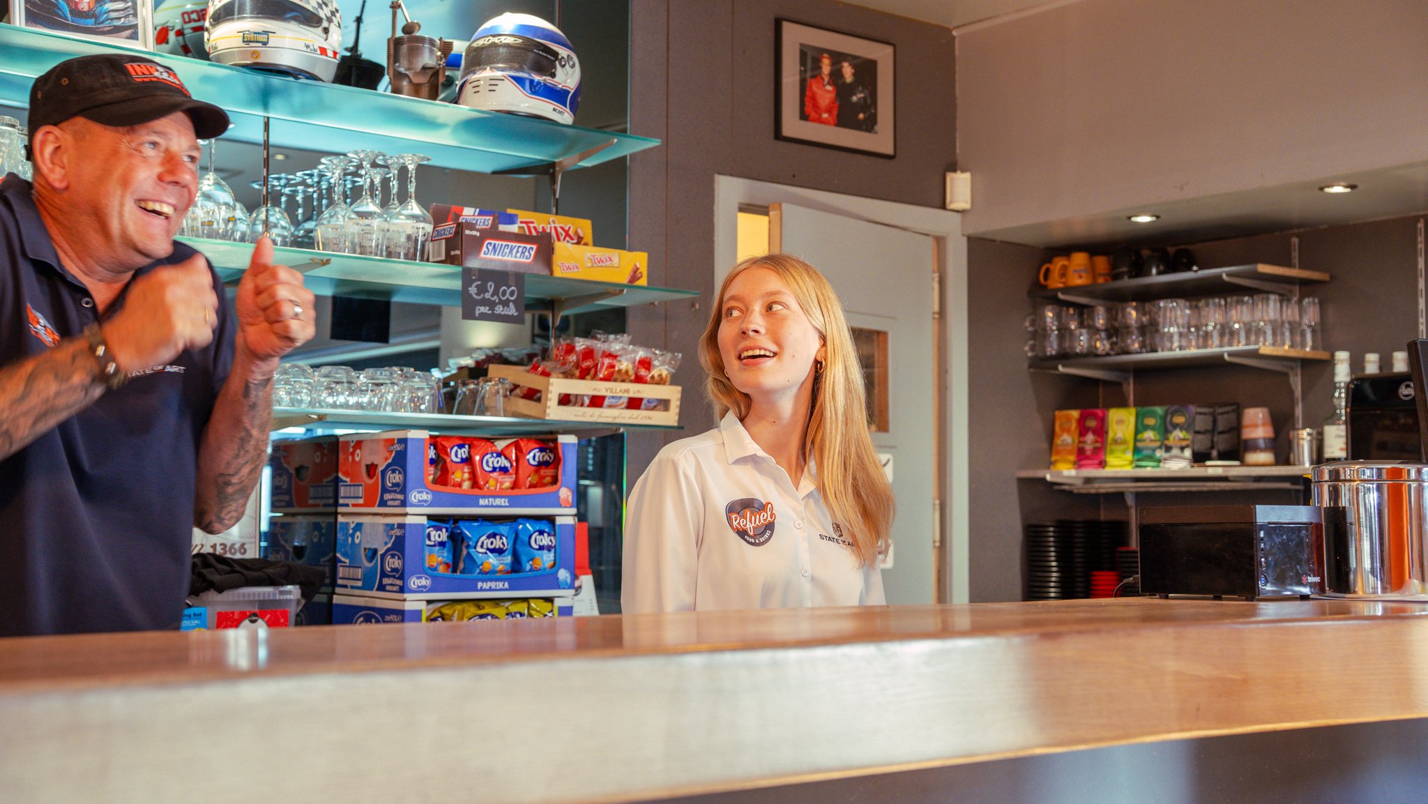 A man and a young woman are behind a counter at a snack bar or café, smiling and engaged in a conversation. The man is wearing a dark polo shirt and cap, and the woman is dressed in a white shirt with a name tag. Behind them are shelves stocked with 