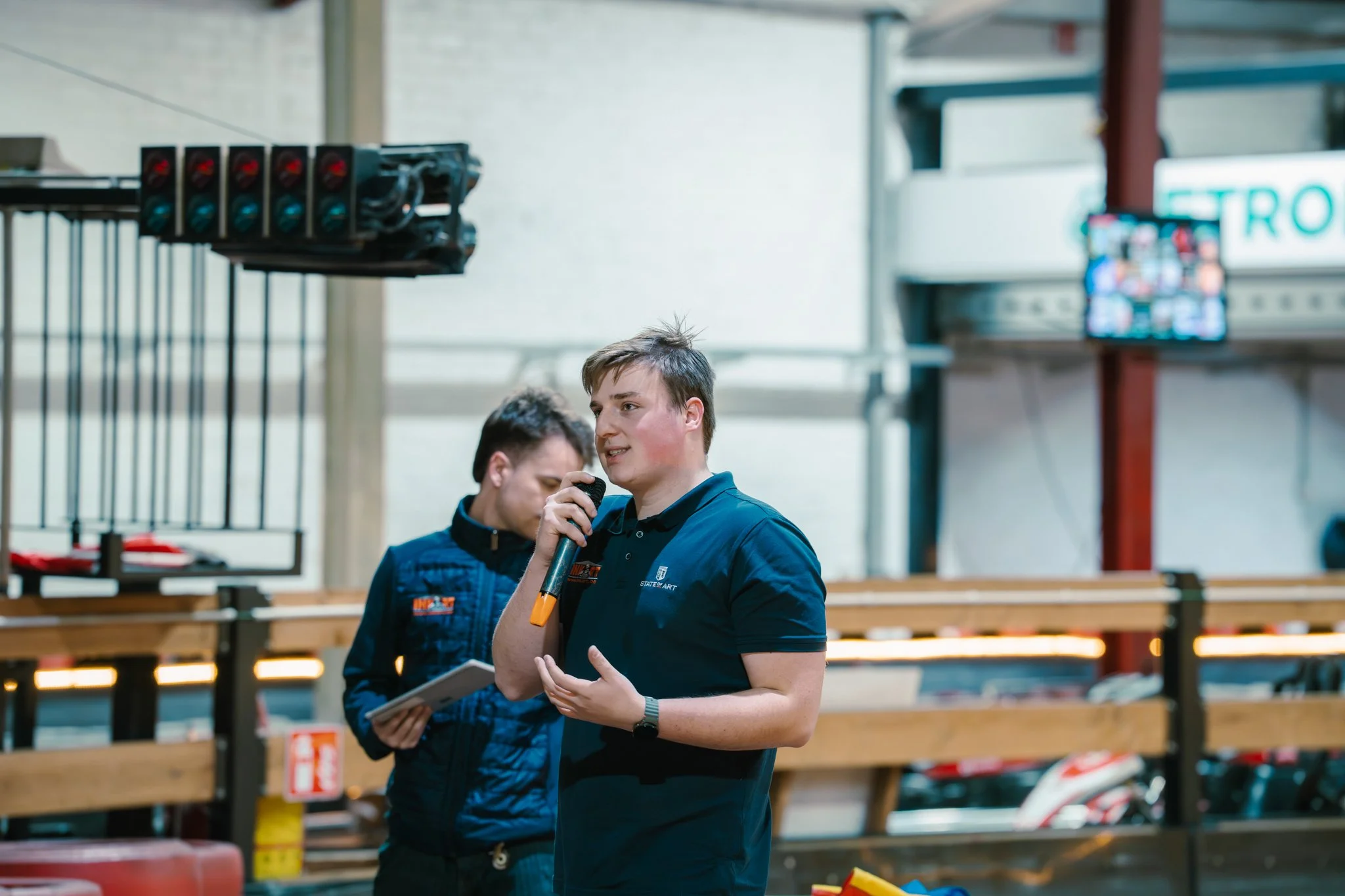 Two young men, one holding a microphone, standing in an indoor sports venue near racing helmets on a shelf, with a scoreboard and electronic screen in the background.