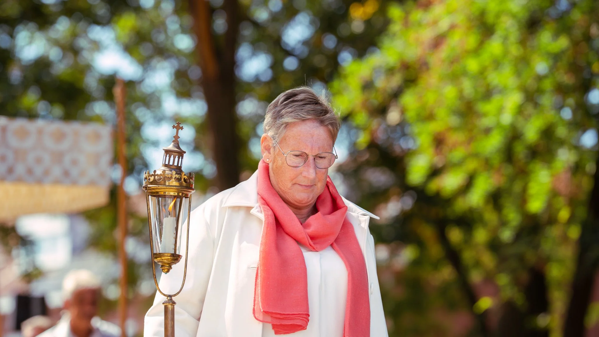 A woman with short hair, glasses, wearing a white jacket and a pink scarf, standing outdoors. There is a decorative lantern with a candle attached to a pole nearby. The background features green foliage and blurred figures.