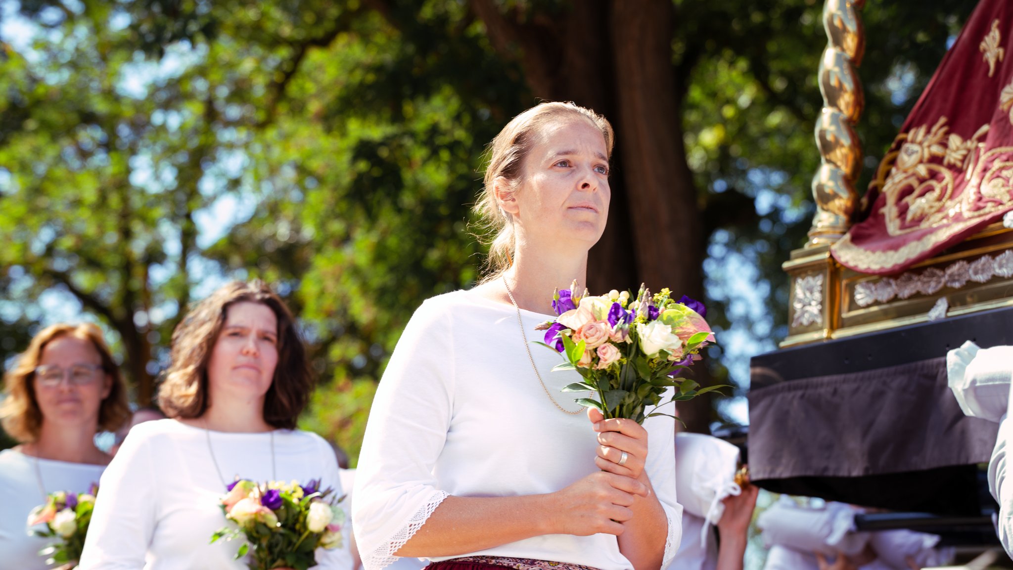 Women dressed in white participating in a religious or cultural procession outdoors, holding bouquets of flowers, with a large decorated religious float nearby, surrounded by green trees.