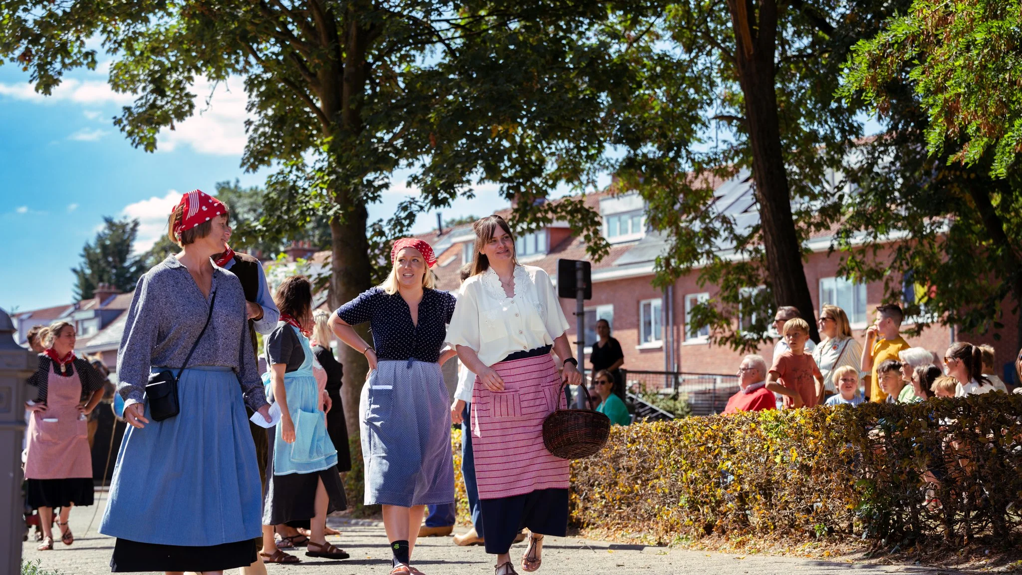 Women dressed in vintage clothing and headscarves walking outdoors under trees, with a group of children and adults in the background, during daytime with a partly cloudy sky.