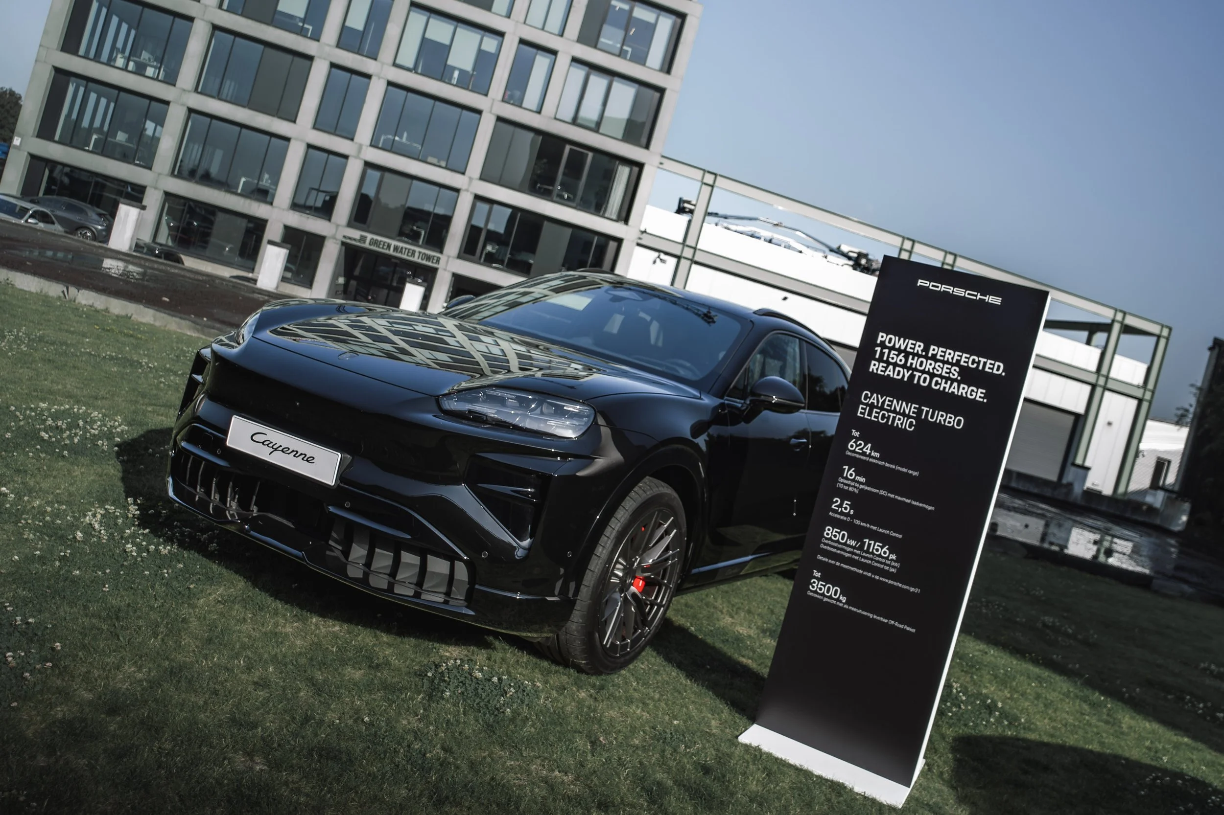 A black Porsche Cayenne Turbo electric vehicle is parked on a grassy area next to a modern building. A large informational sign displays specifications about the vehicle, including power, range, and charging details.
