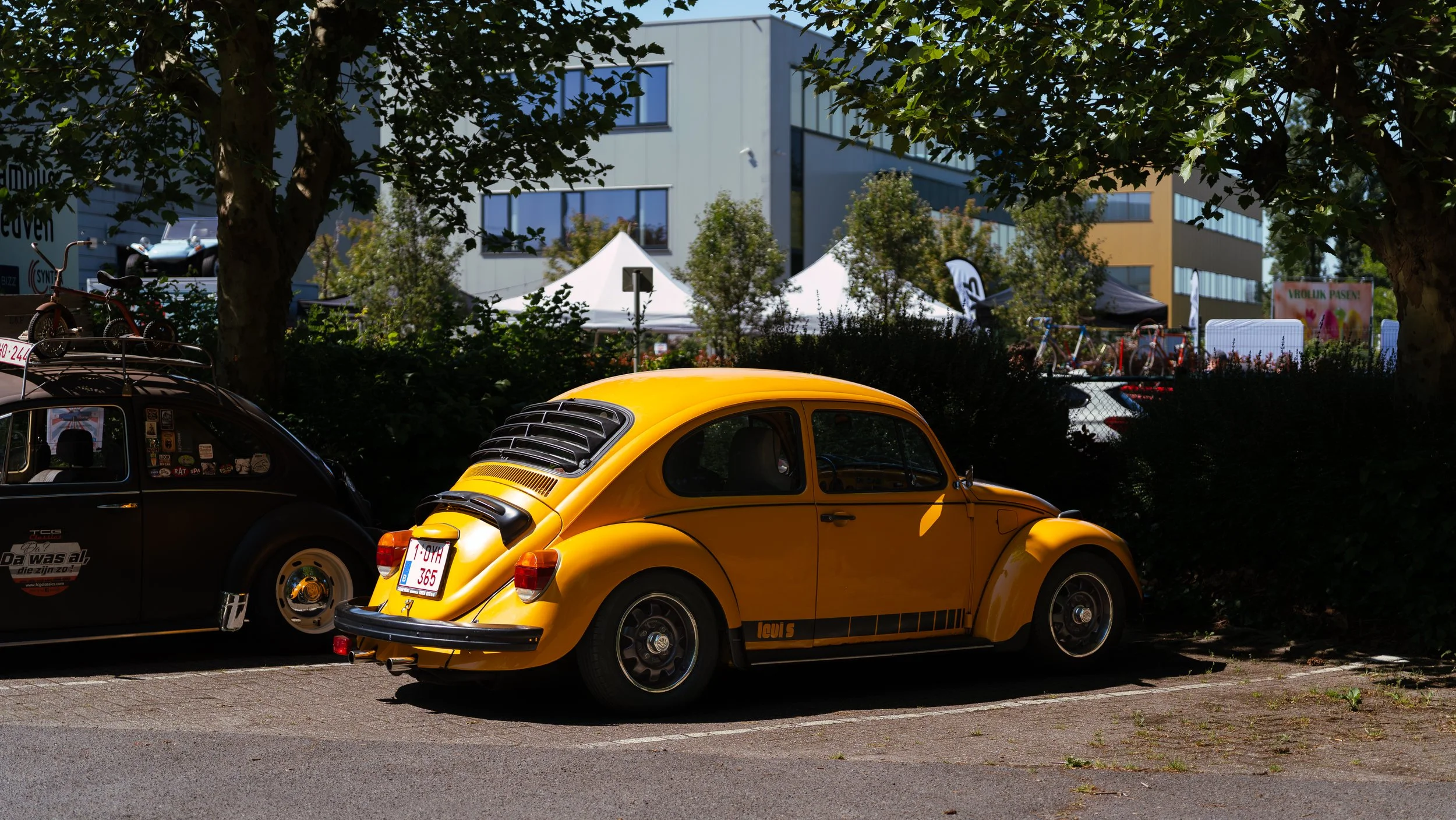 Yellow vintage Volkswagen Beetle parked in a parking lot beside a black car, with trees and modern buildings in the background.
