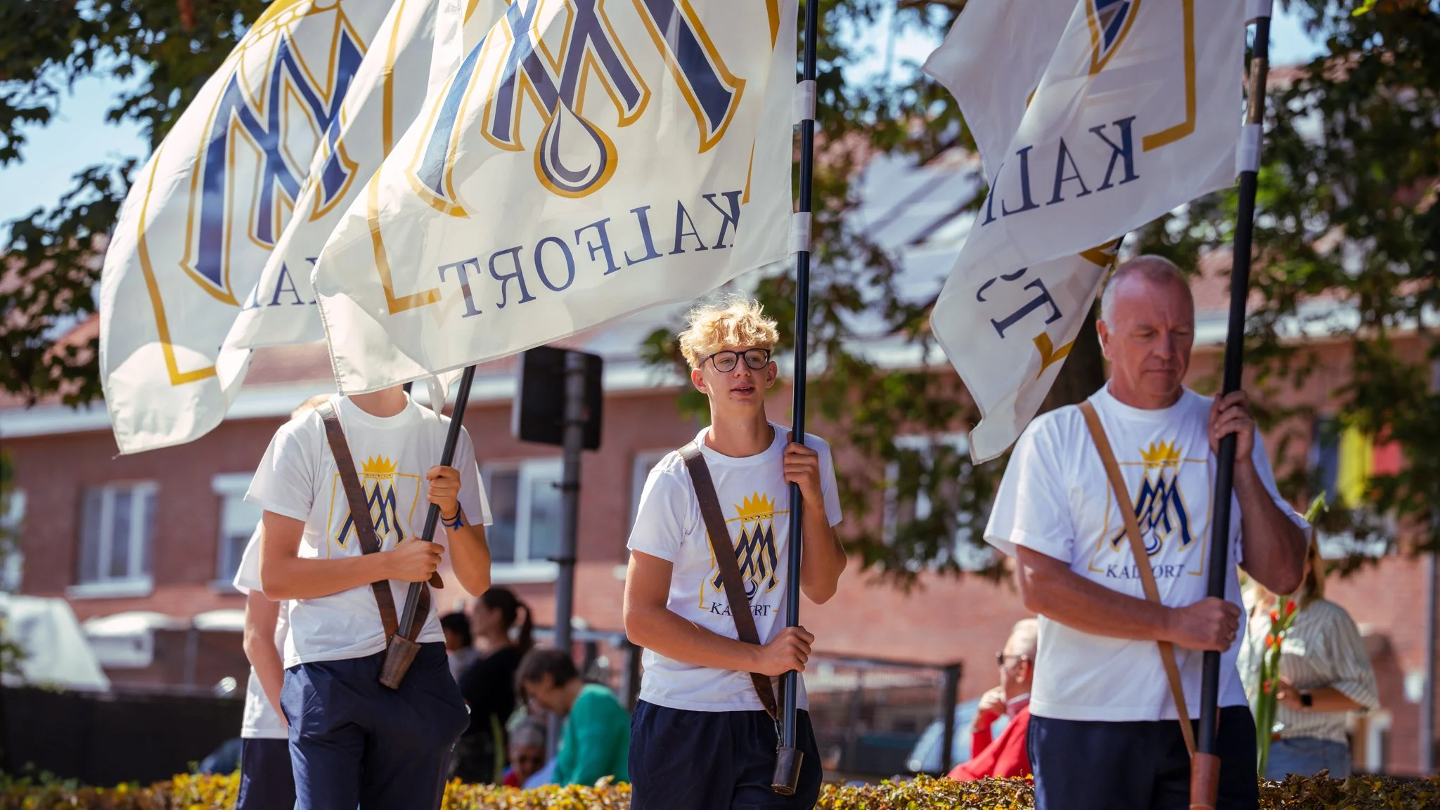 People participating in a parade or rally, carrying flags with a crown and M logo, dressed in matching white T-shirts with a similar logo, outdoors with trees and buildings in the background.