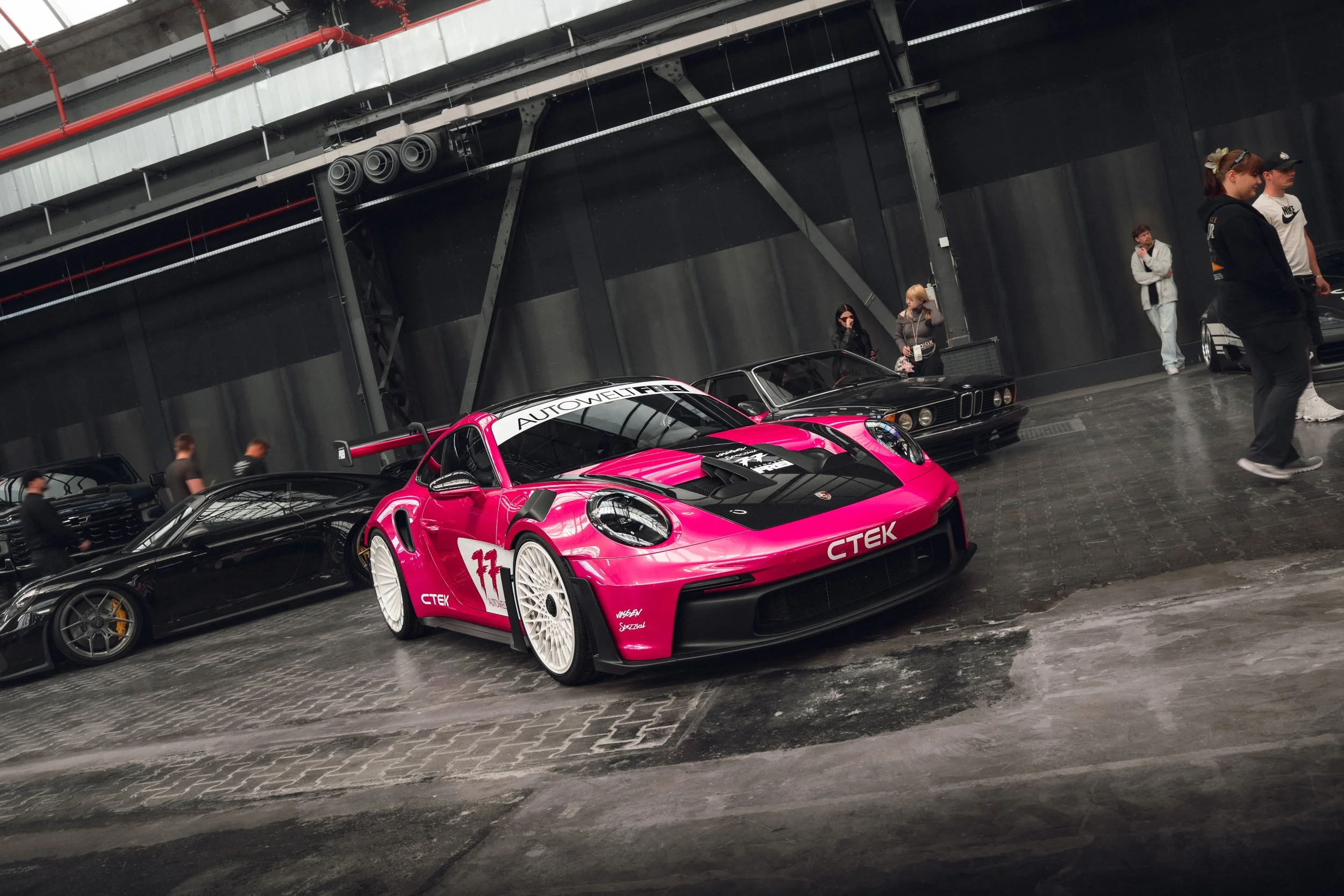A pink race car with a black hood and white wheels in an indoor exhibition space. Other cars and people are visible in the background.
