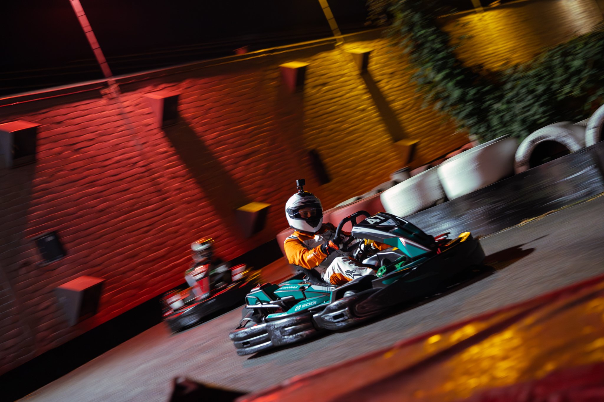 Two people racing go-karts on an indoor track at night, with a brick wall in the background illuminated by red, yellow, and green lights, and barriers along the track.