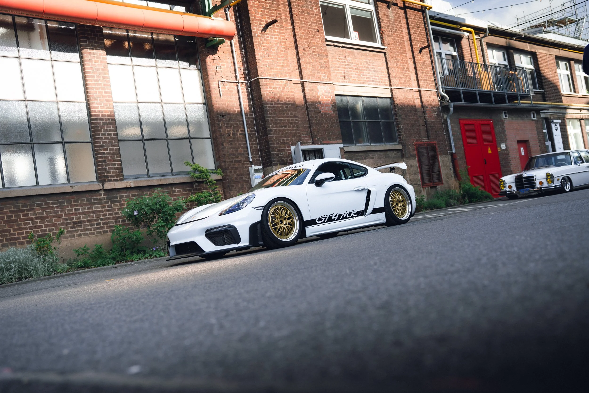 A white sports car with gold rims and a spoiler parked on the street in front of a brick building with large windows, and another classic white car parked nearby. The building has red and yellow pipes and a small balcony.