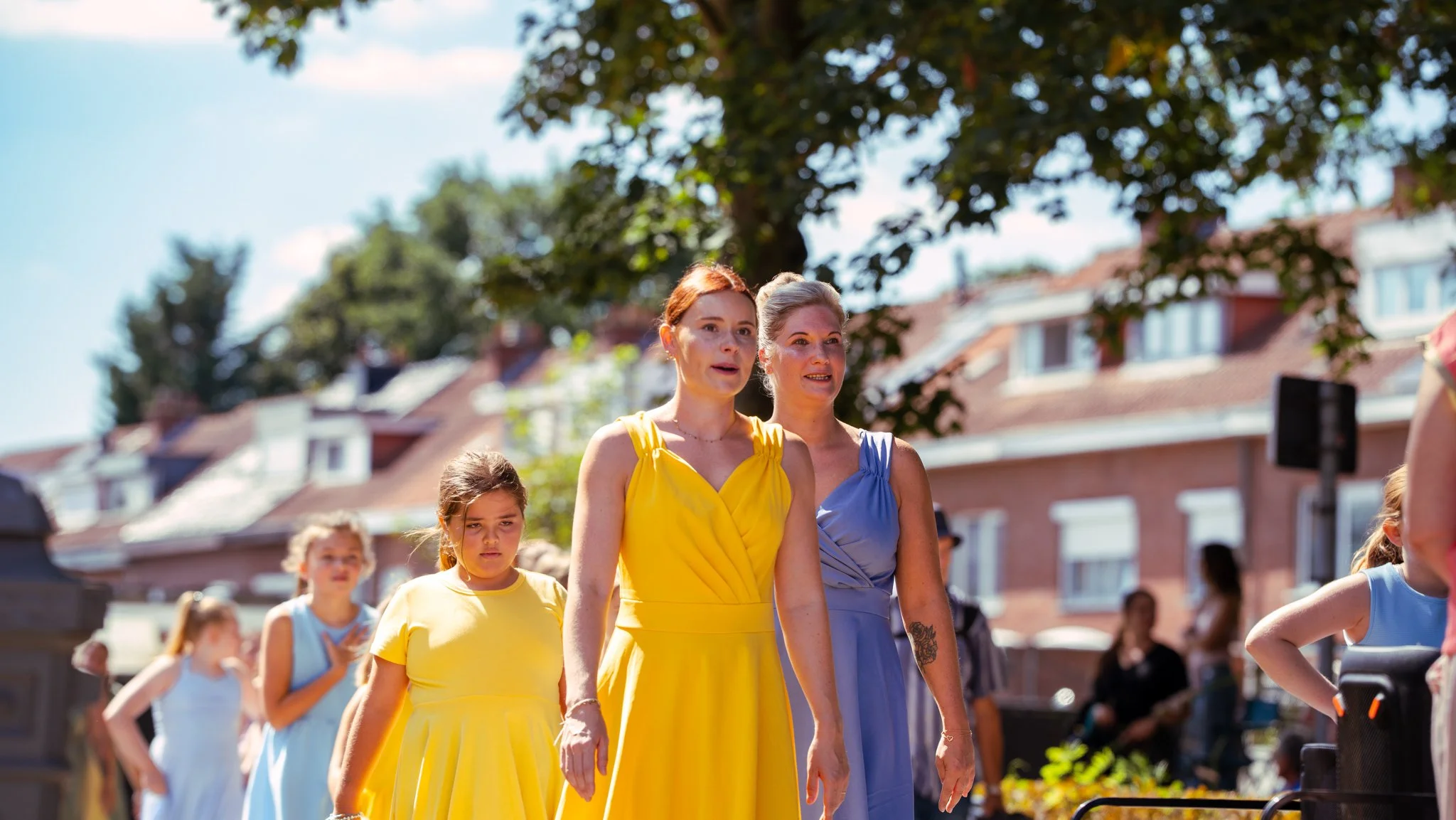 Women and girls in colorful dresses walking outdoors on a sunny day with trees and houses in the background.