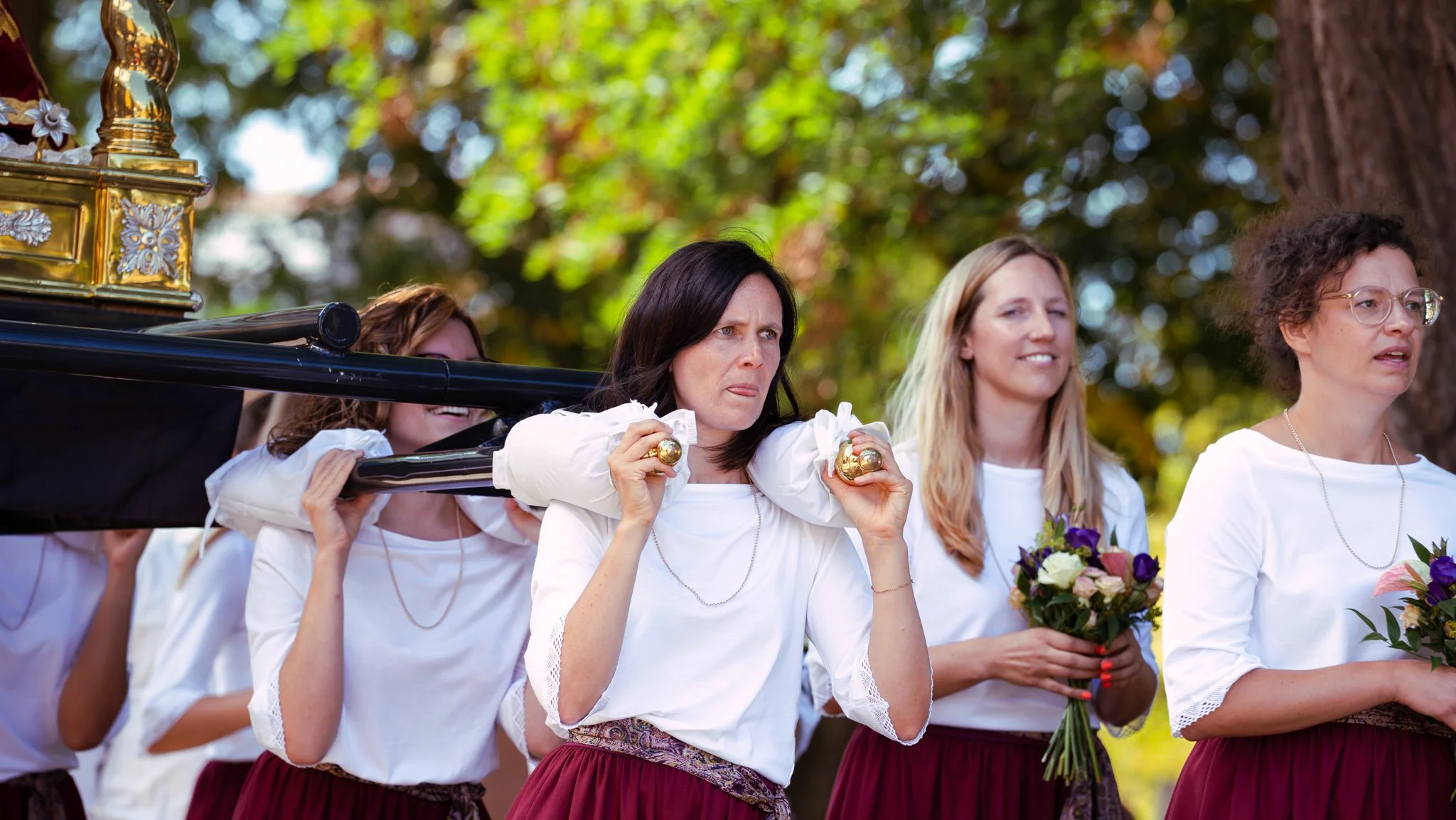 A group of women dressed in white blouses and maroon skirts participate in an outdoor cultural event, carrying a decorated float.