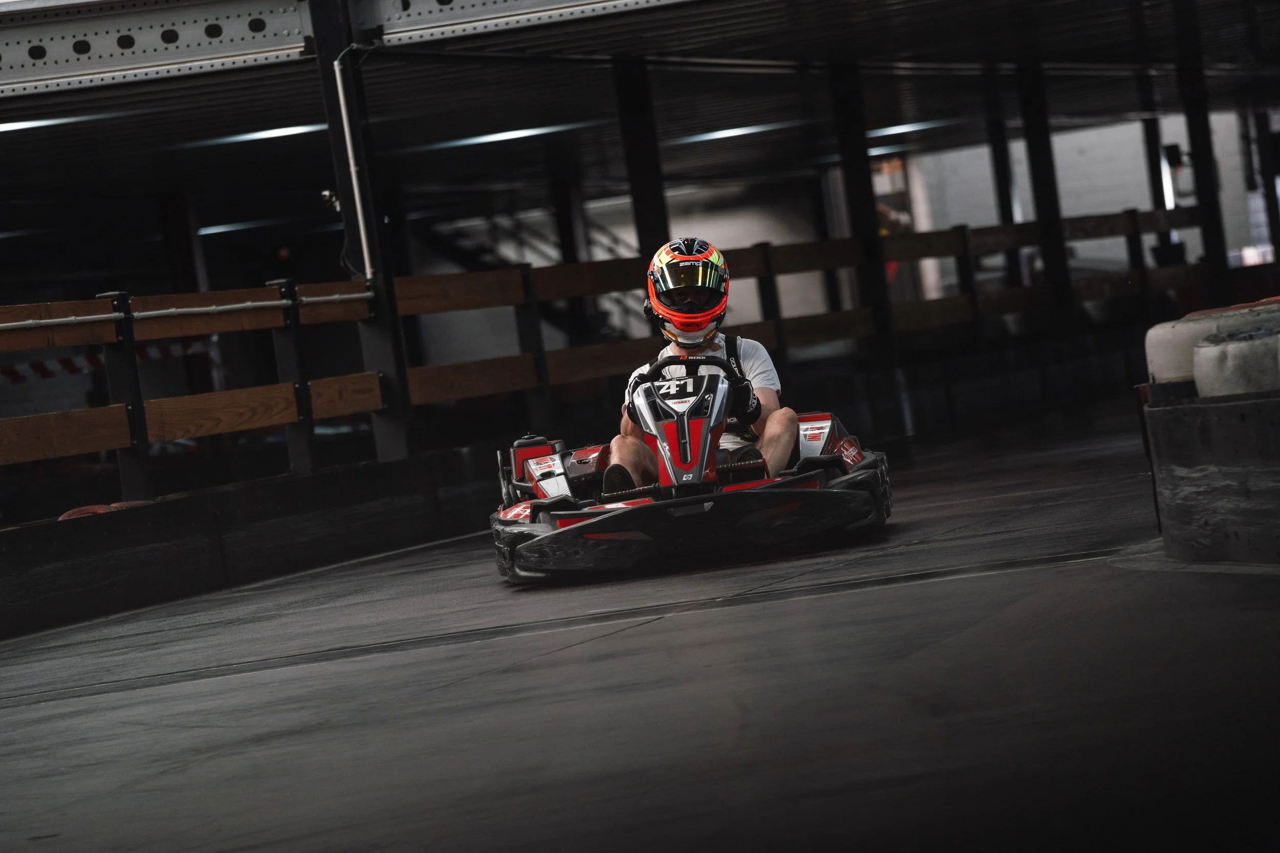 A person wearing a helmet is driving a go-kart on an indoor track, with blurred surroundings and barriers on the sides.