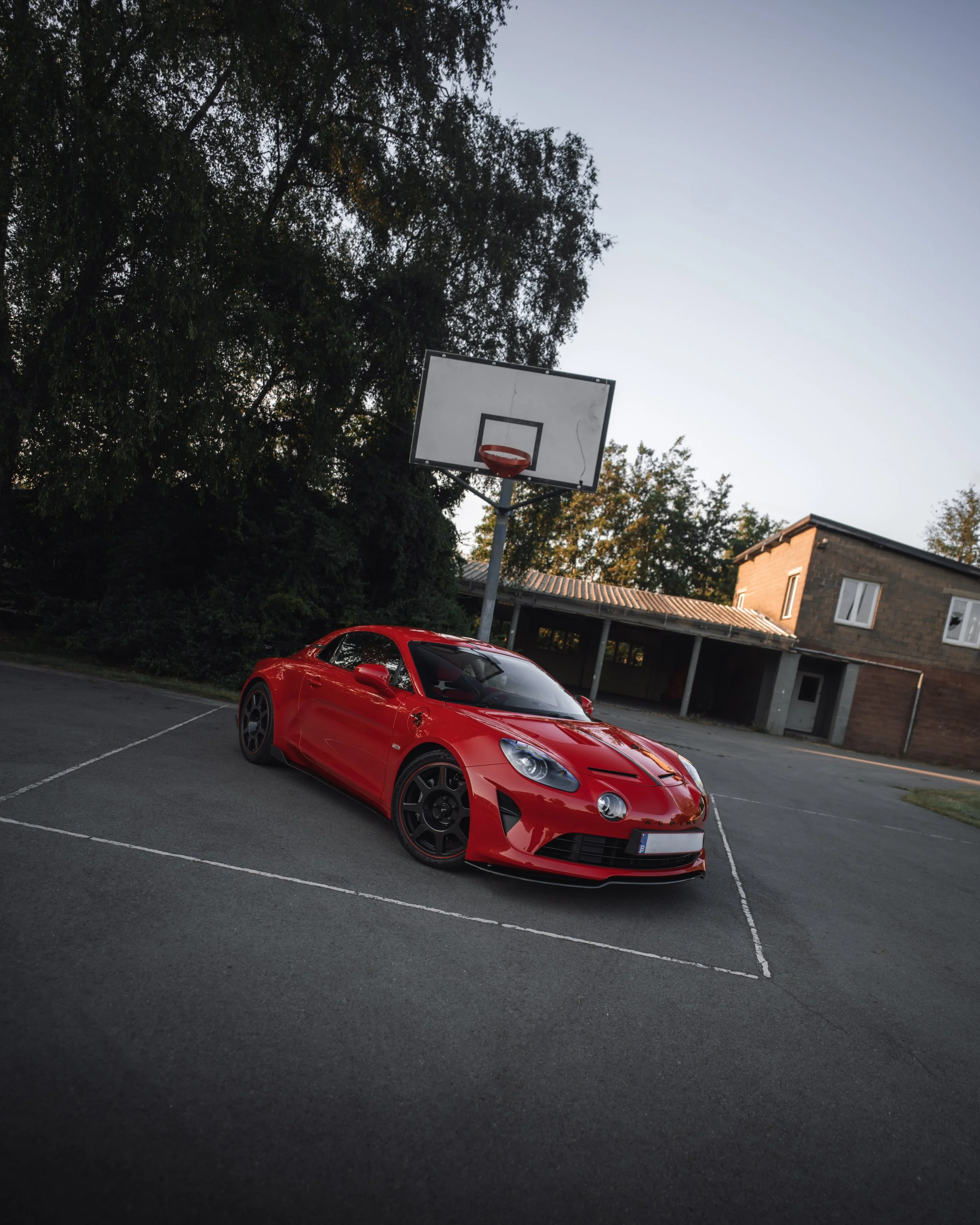 Red sports car parked on a marked parking spot near a basketball hoop under a tree with a building in the background.