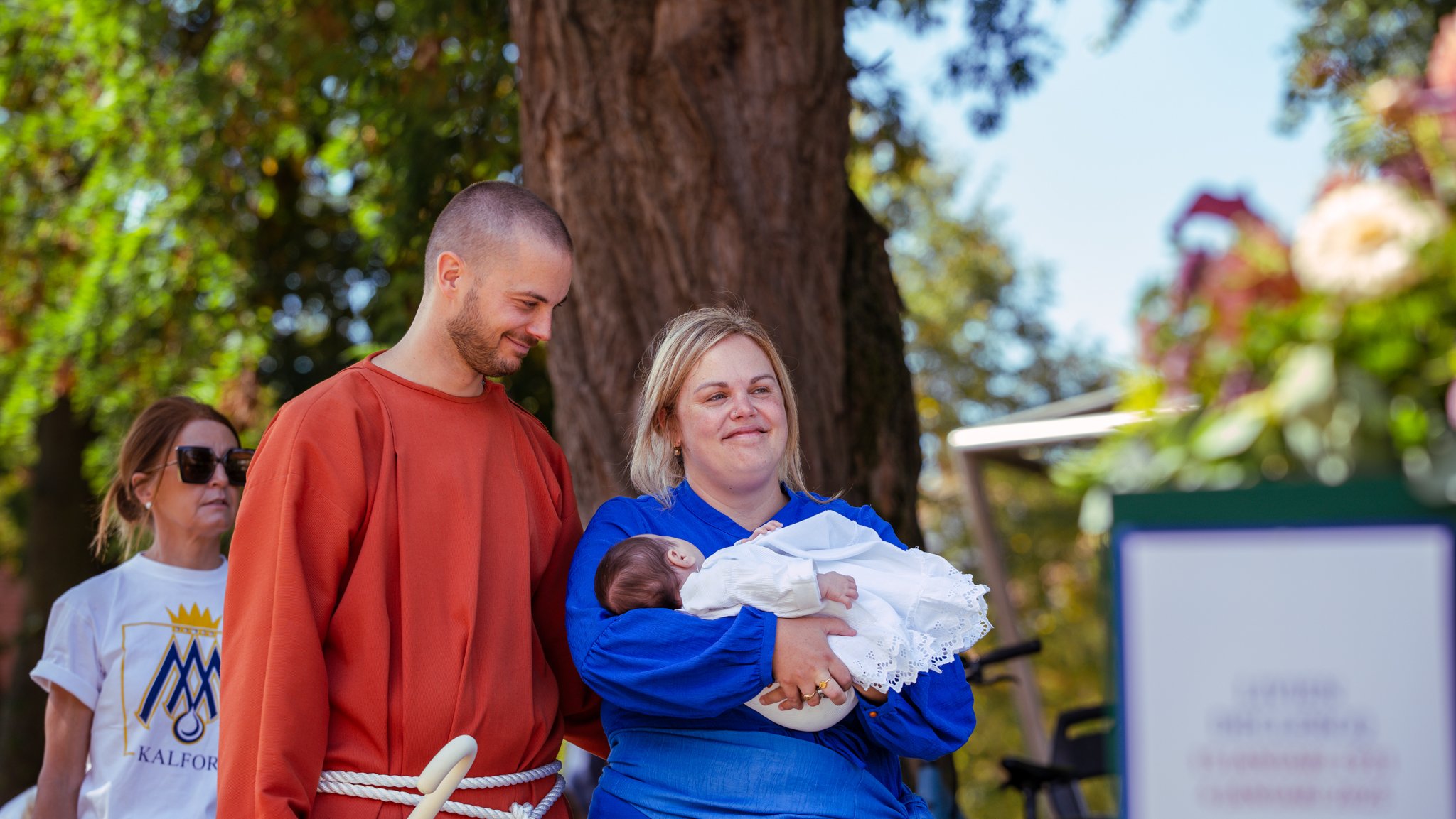 A woman holding a newborn baby in her arms while standing outdoors near a tree during daytime, with a man standing beside her looking down at the baby and a woman in sunglasses in the background. The woman with the baby is smiling, and the scene appe