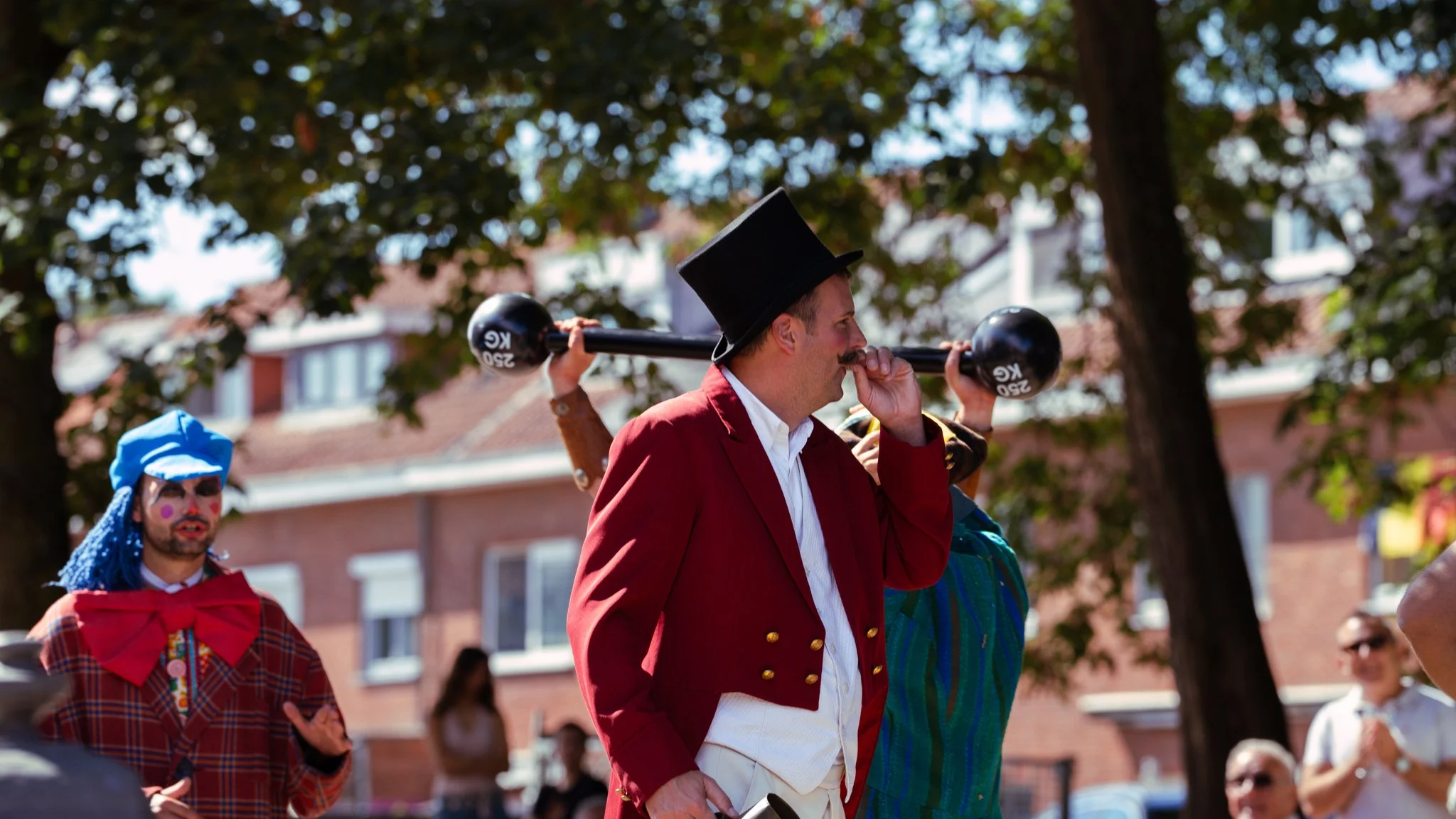 A man dressed as Abraham Lincoln in a red coat and top hat carries a black sledgehammer over his shoulder during a street parade or festival, with other costumed individuals and spectators in the background.