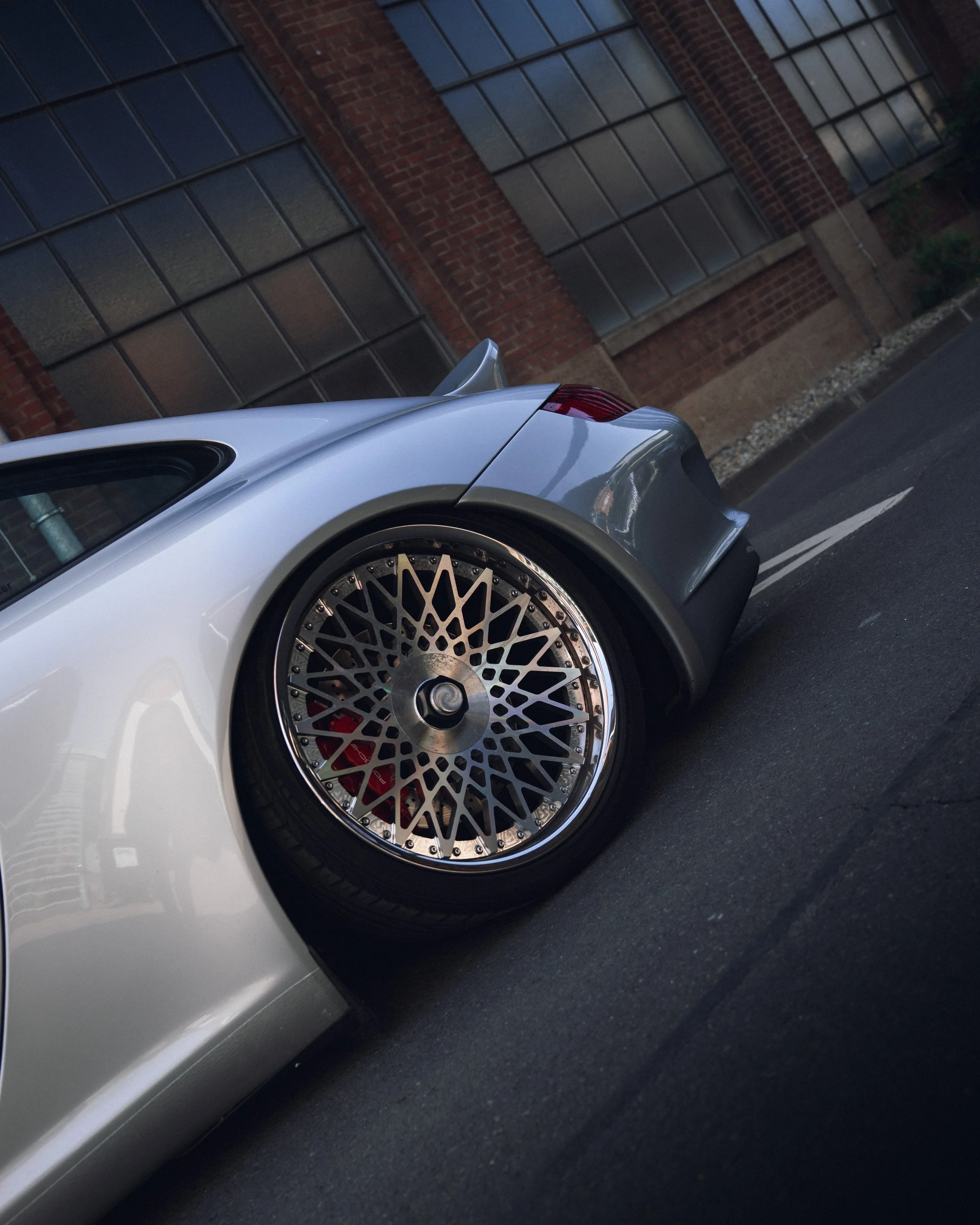 Close-up view of the rear end of a silver sports car, focusing on the intricate, multi-spoke alloy wheel and tire, with a background of brick building with large windows.