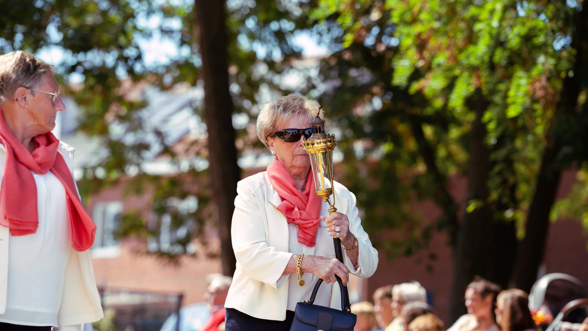 An elderly woman with sunglasses and a coral scarf holding a torch during an outdoor event with trees and buildings in the background.