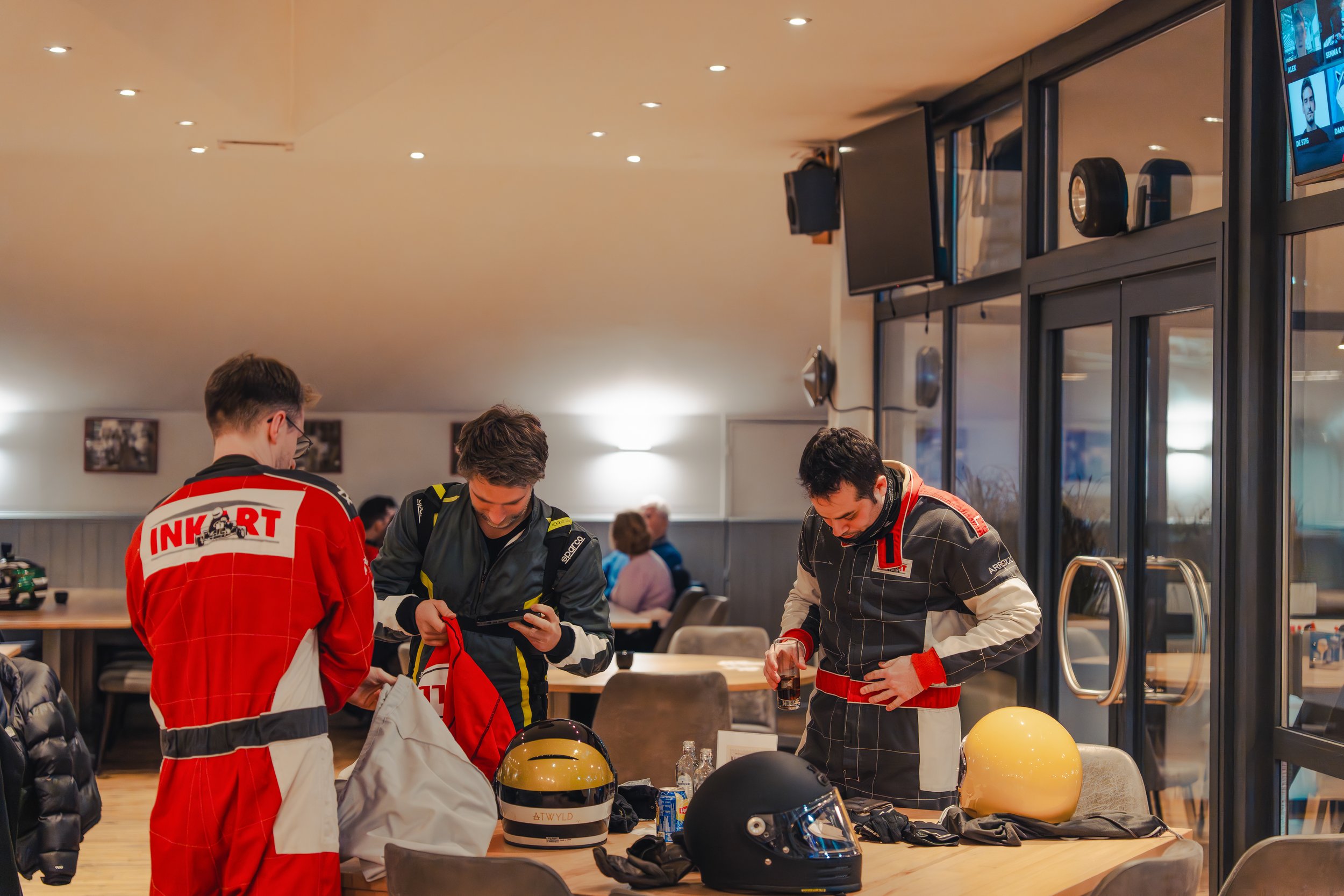 Three people wearing racing suits standing at a table with helmets and gear, inside a room with large glass windows and a TV screen.