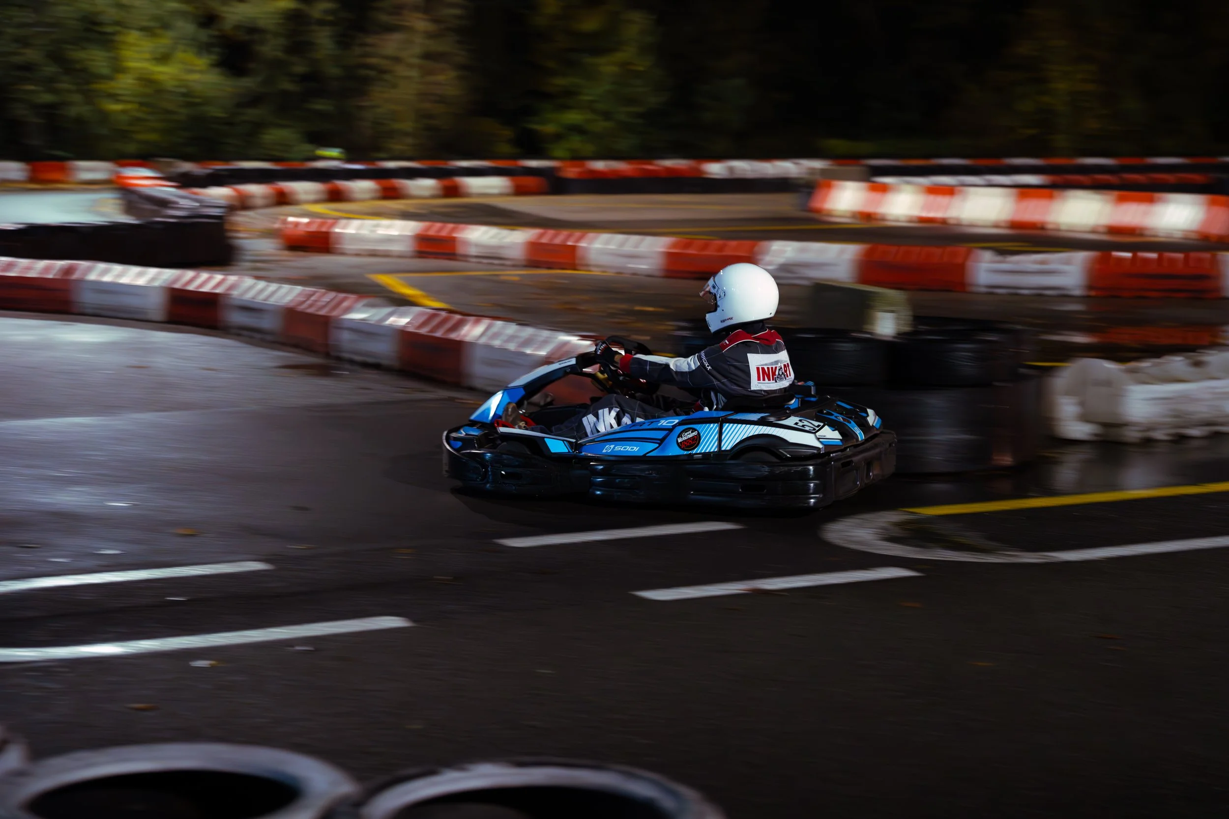 Person in a racing helmet and racing suit driving a go-kart on a sharp turn on an outdoor race track, surrounded by safety barriers.