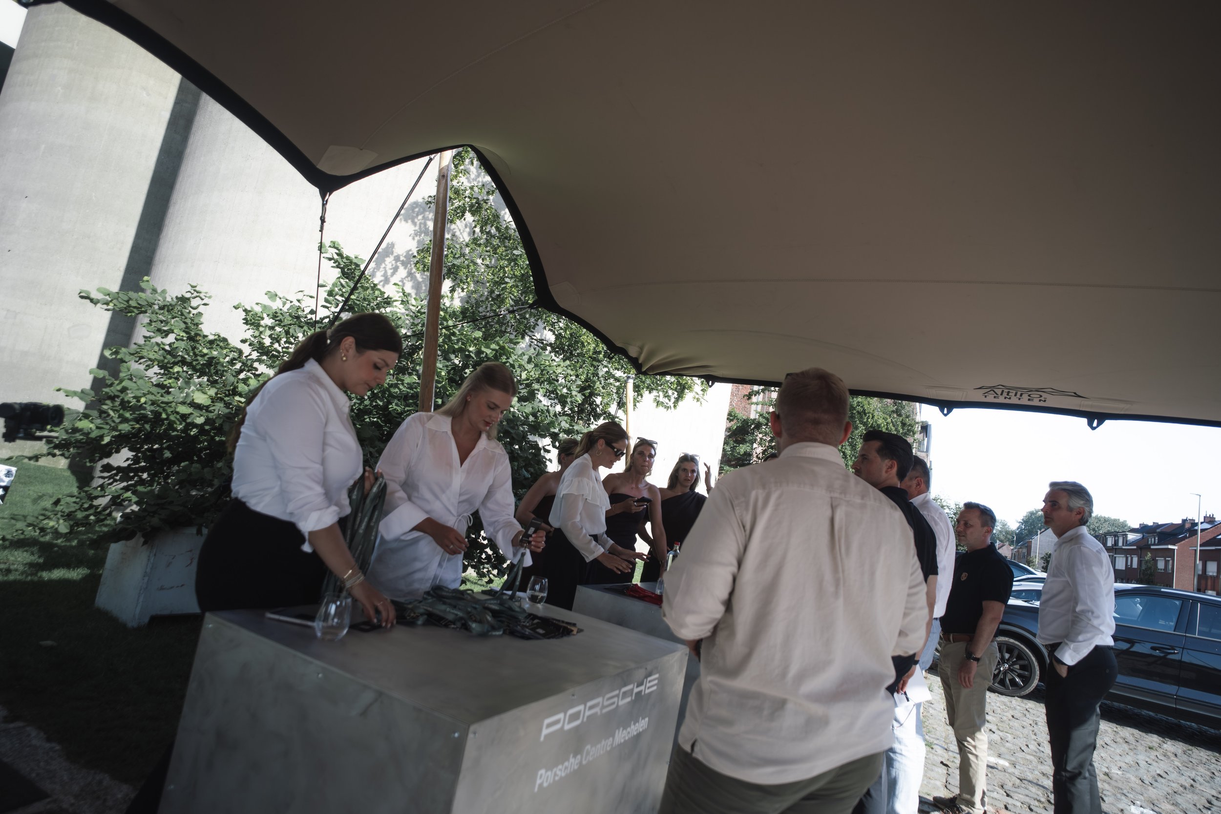 Group of people gathered under a canopy at an outdoor event, some wearing white shirts, with a Porsche sign visible on a table, and cars parked in the background.