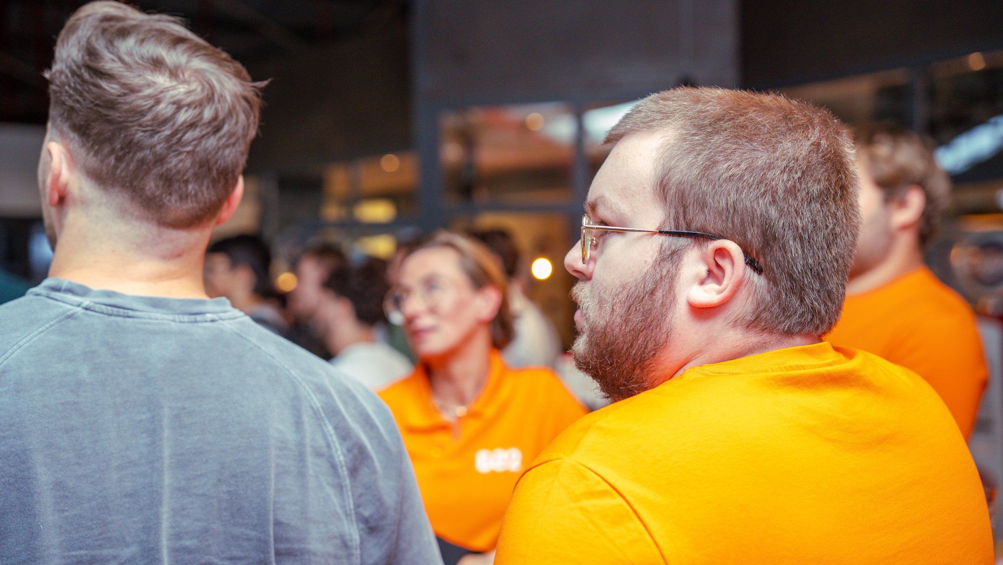 People gathered indoors, some wearing orange shirts, engaged in conversation and socializing.