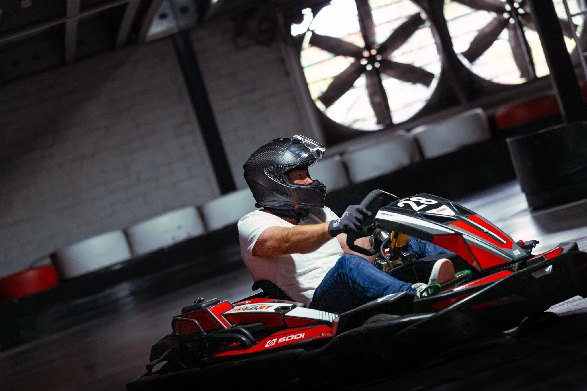 A person wearing a black helmet, white shirt, and black gloves driving a red go-kart indoor race track.