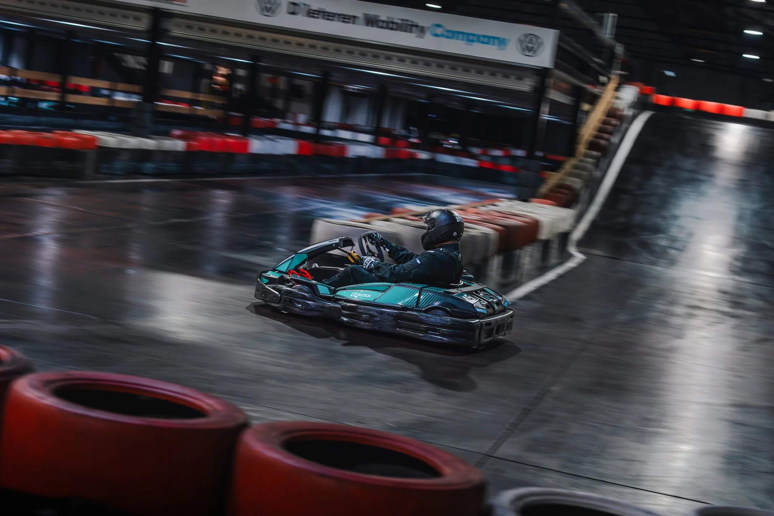 A go-kart racer wearing a black helmet and racing suit navigating a turn on an indoor track, with red and white barriers and blurred background.
