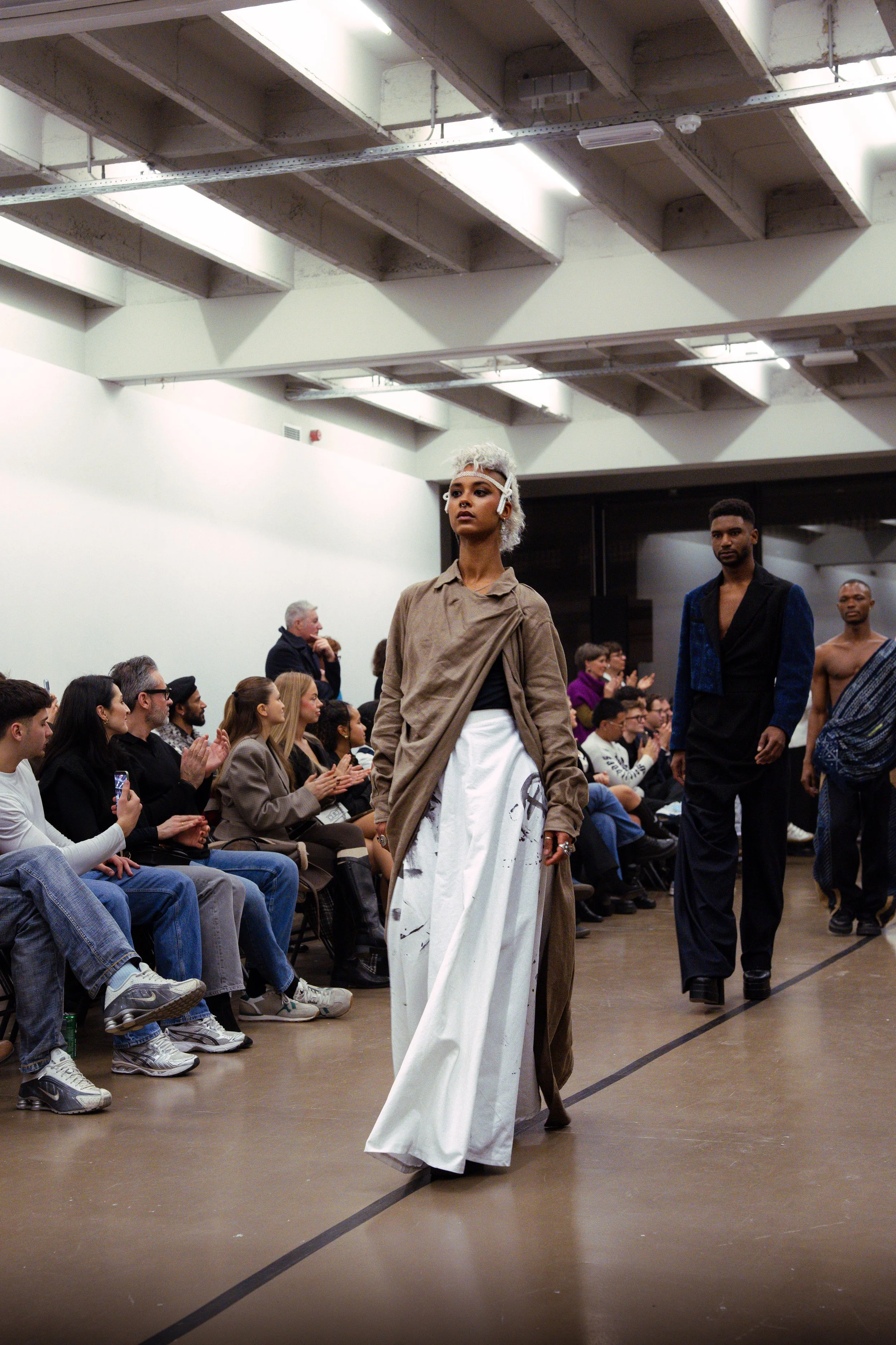 Model walking down runway at a fashion show, audience seated on either side applauding, indoor setting with industrial ceiling, diverse attendees.