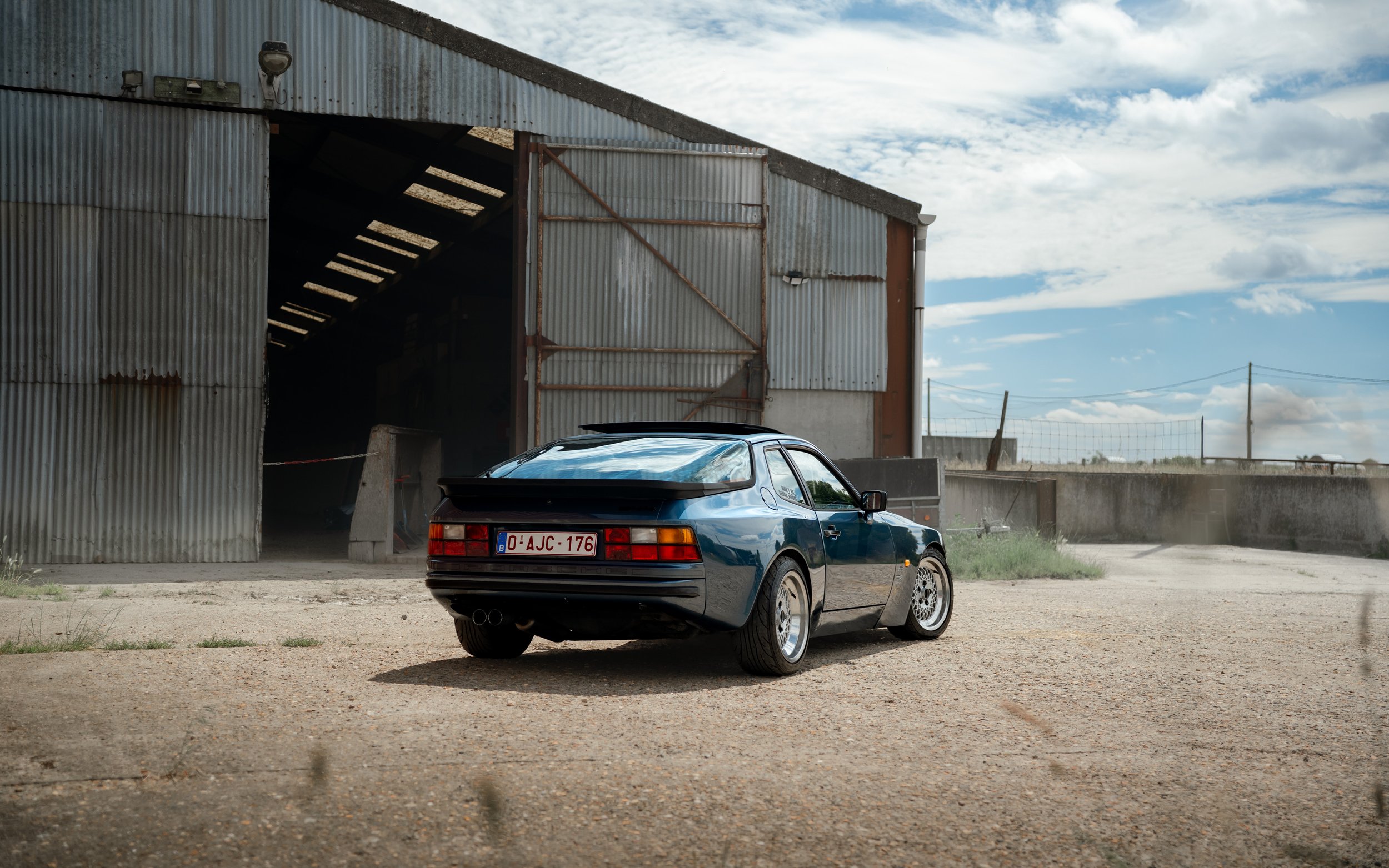 A black sports car with a European license plate parked in front of a large rusted metal barn with a staircase inside, set against a partly cloudy sky.