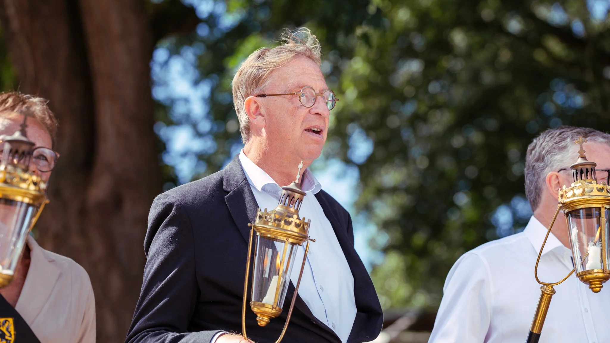 Three men wearing white shirts and black jackets holding lanterns during a daytime outdoor event.