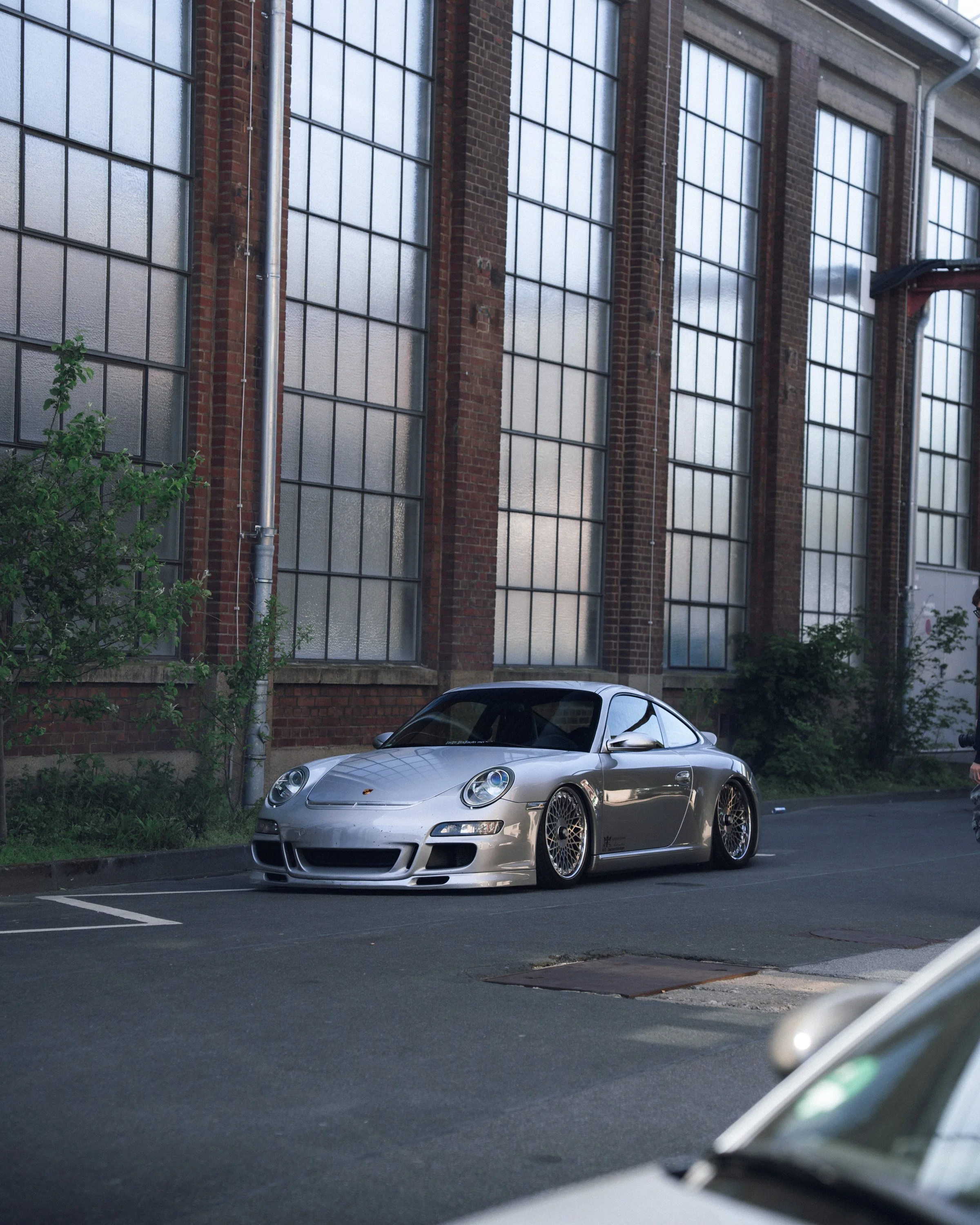 Silver Porsche sports car parked in an urban parking lot in front of a brick building with large windows. The photo appears to be taken in the evening or early morning.