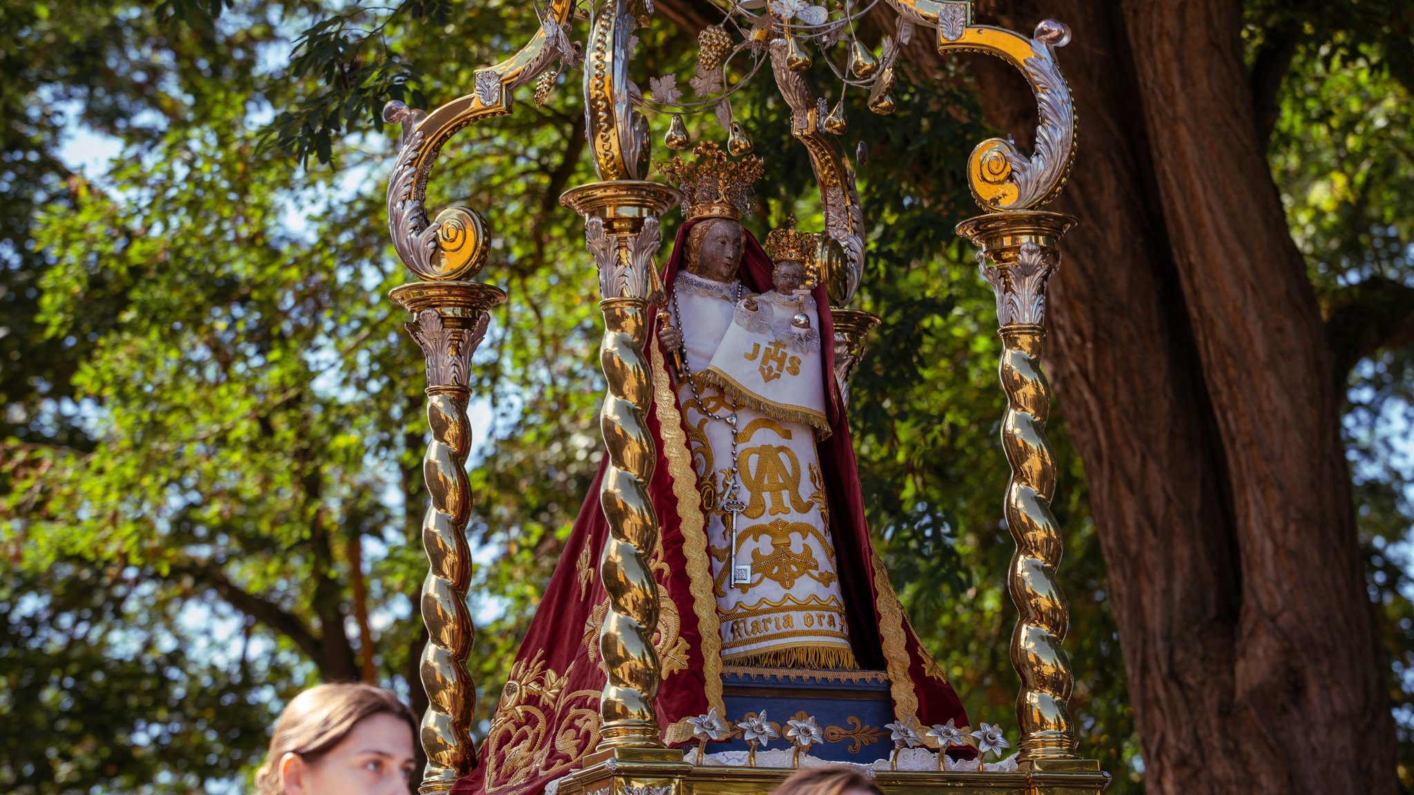 A religious statue of the Virgin Mary holding baby Jesus, adorned with crowns and ornate garments, displayed on a gilded platform with decorative columns during an outdoor celebration near trees.