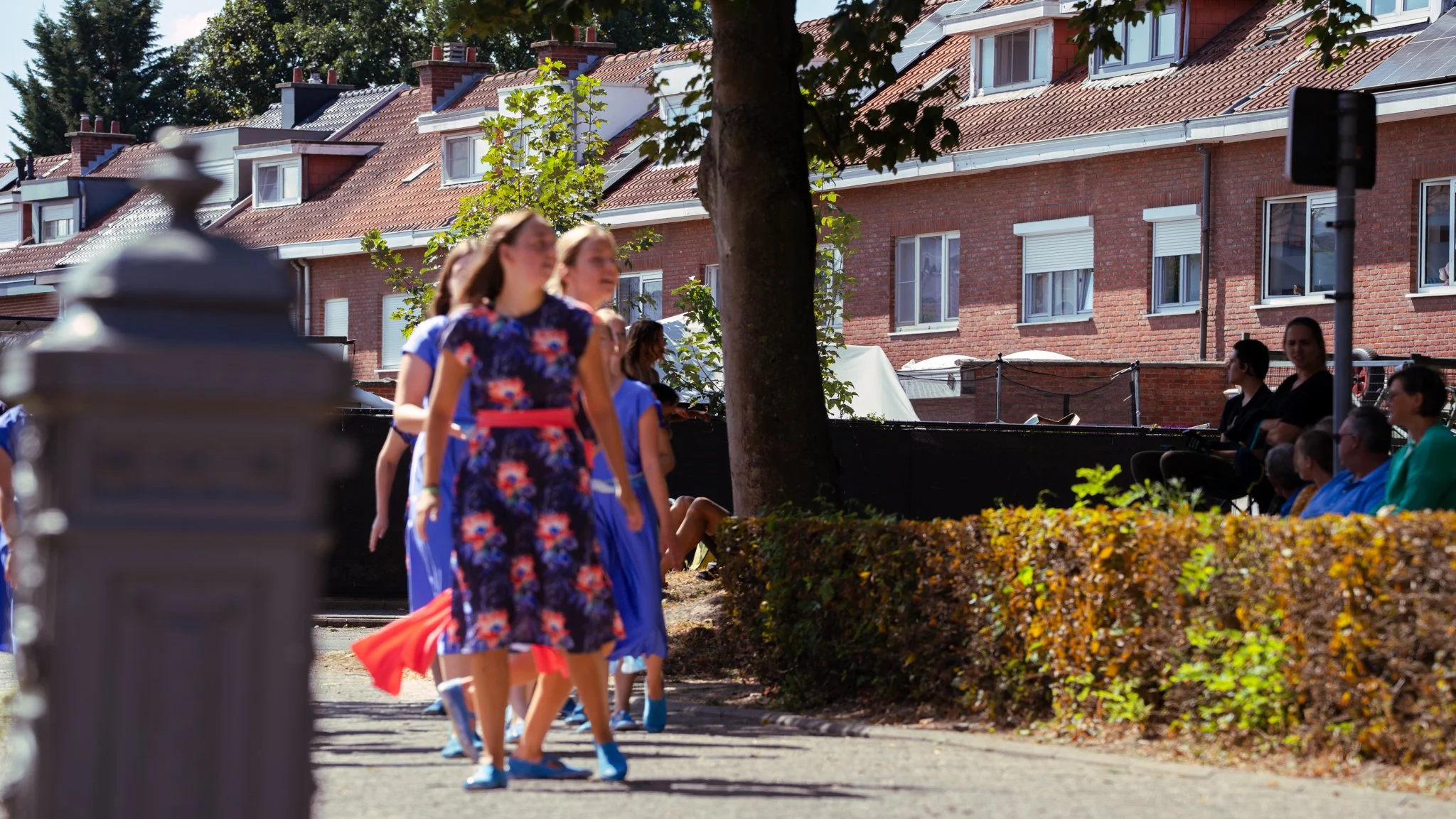 Girls in blue and floral dresses walking along a sidewalk during a parade or festival, with houses and spectators seated on a bench in the background.