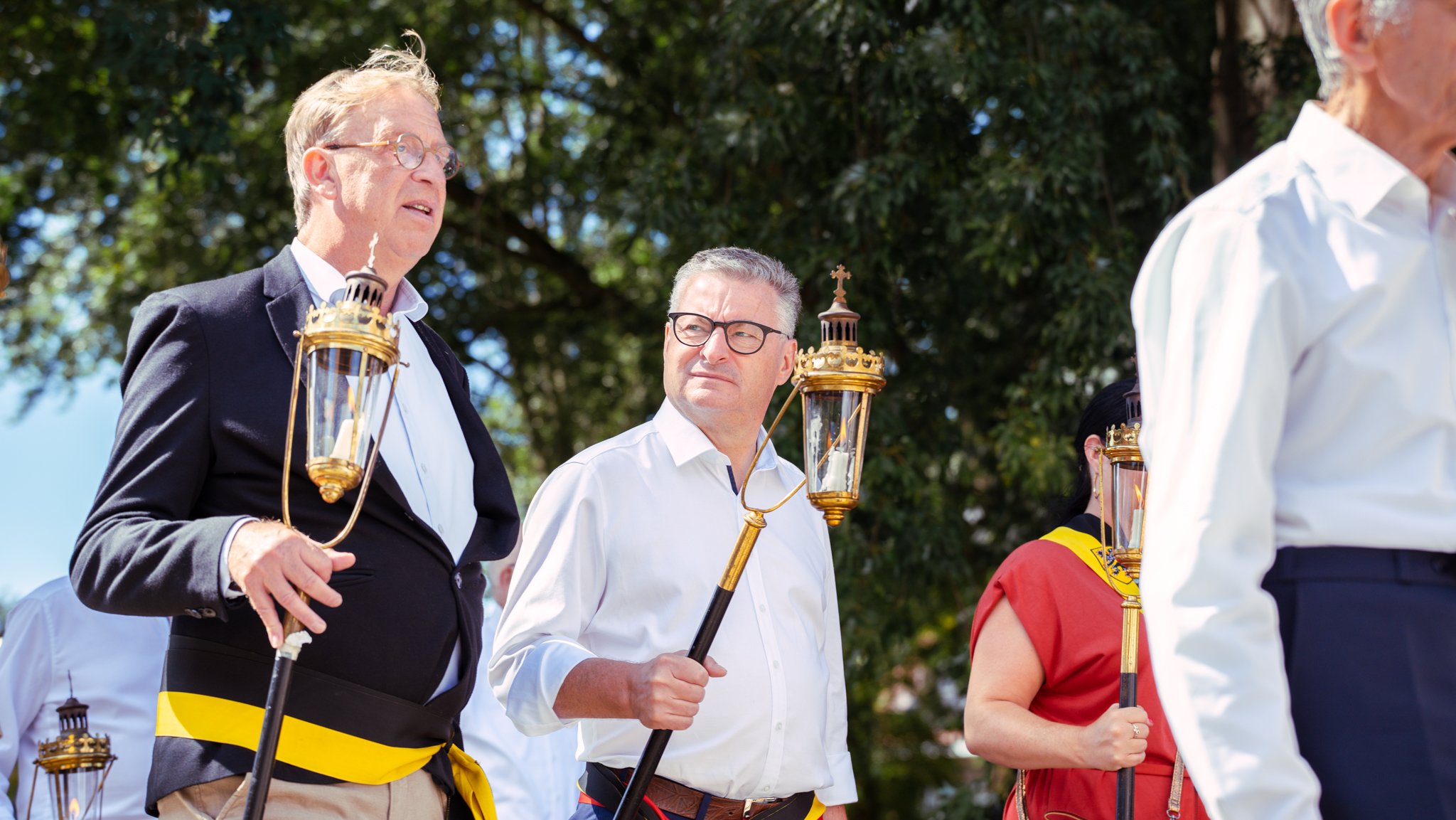Men and women participating in a religious procession holding lanterns with candles, outdoors under trees.