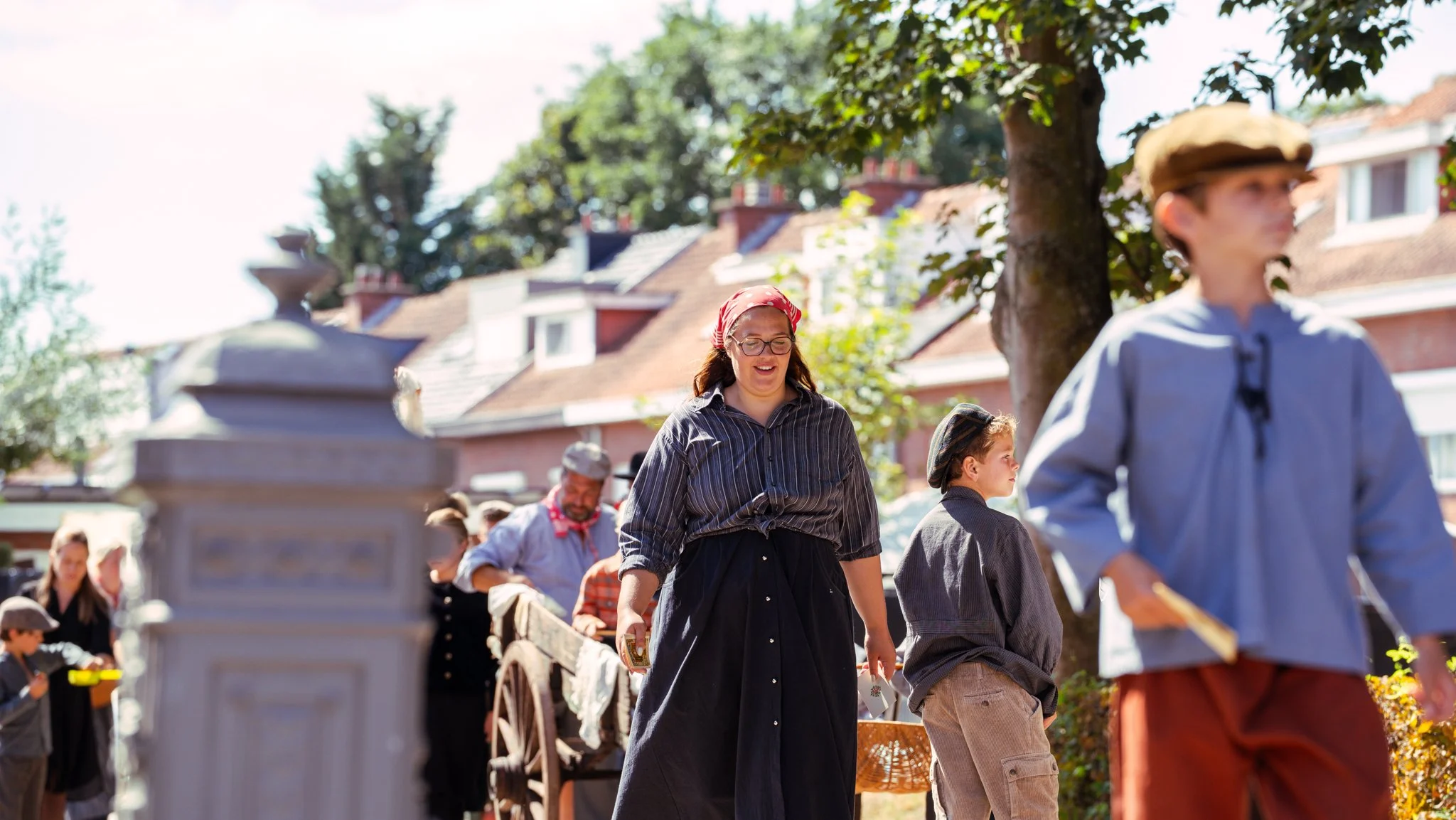 People dressed in vintage clothing, walking outdoors on a sunny day, with striped shirts and hats, near houses and trees.