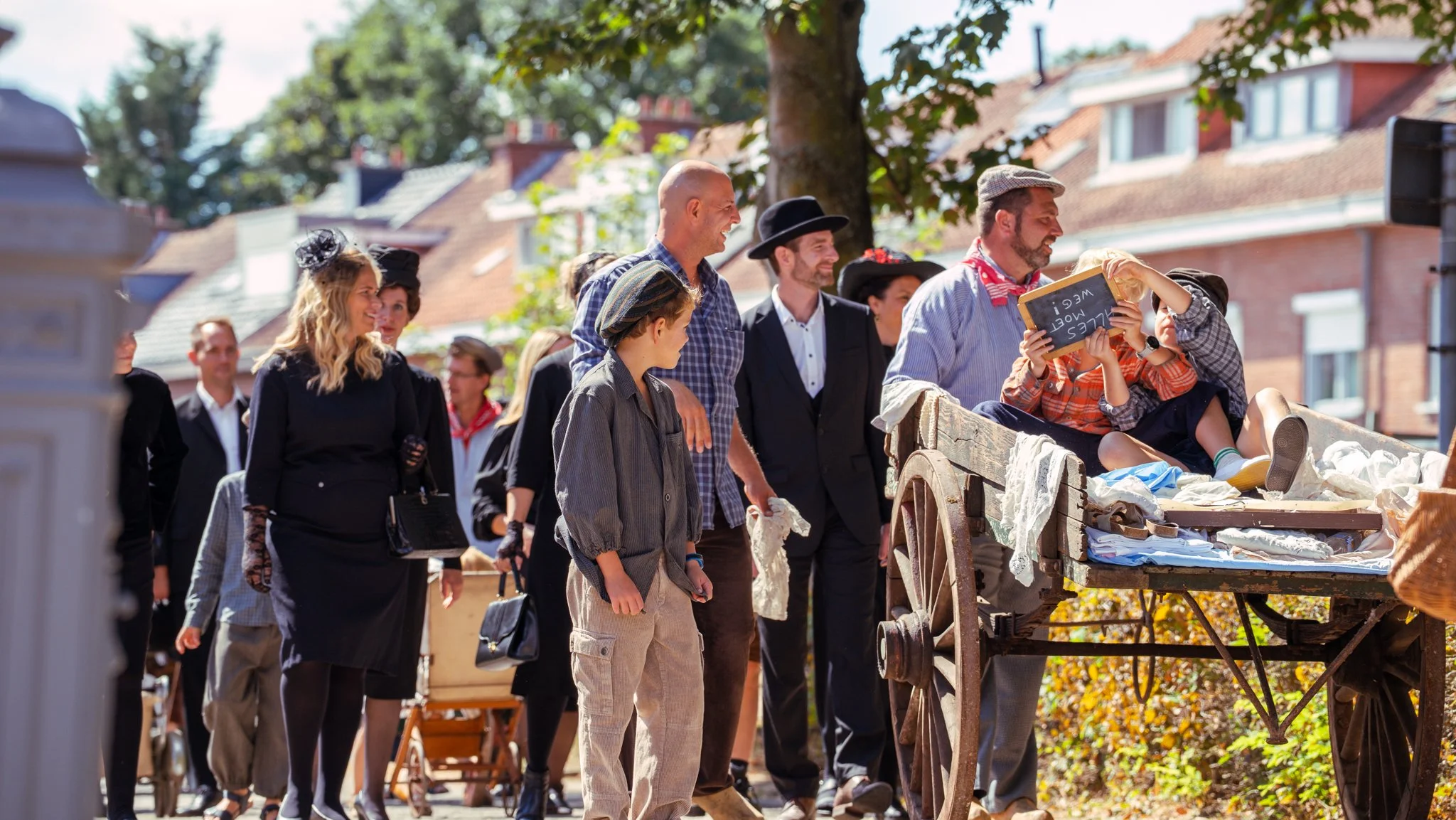 Group of people in vintage clothing at an outdoor market or event, browsing and chatting around a wooden cart with fabrics and items on display
