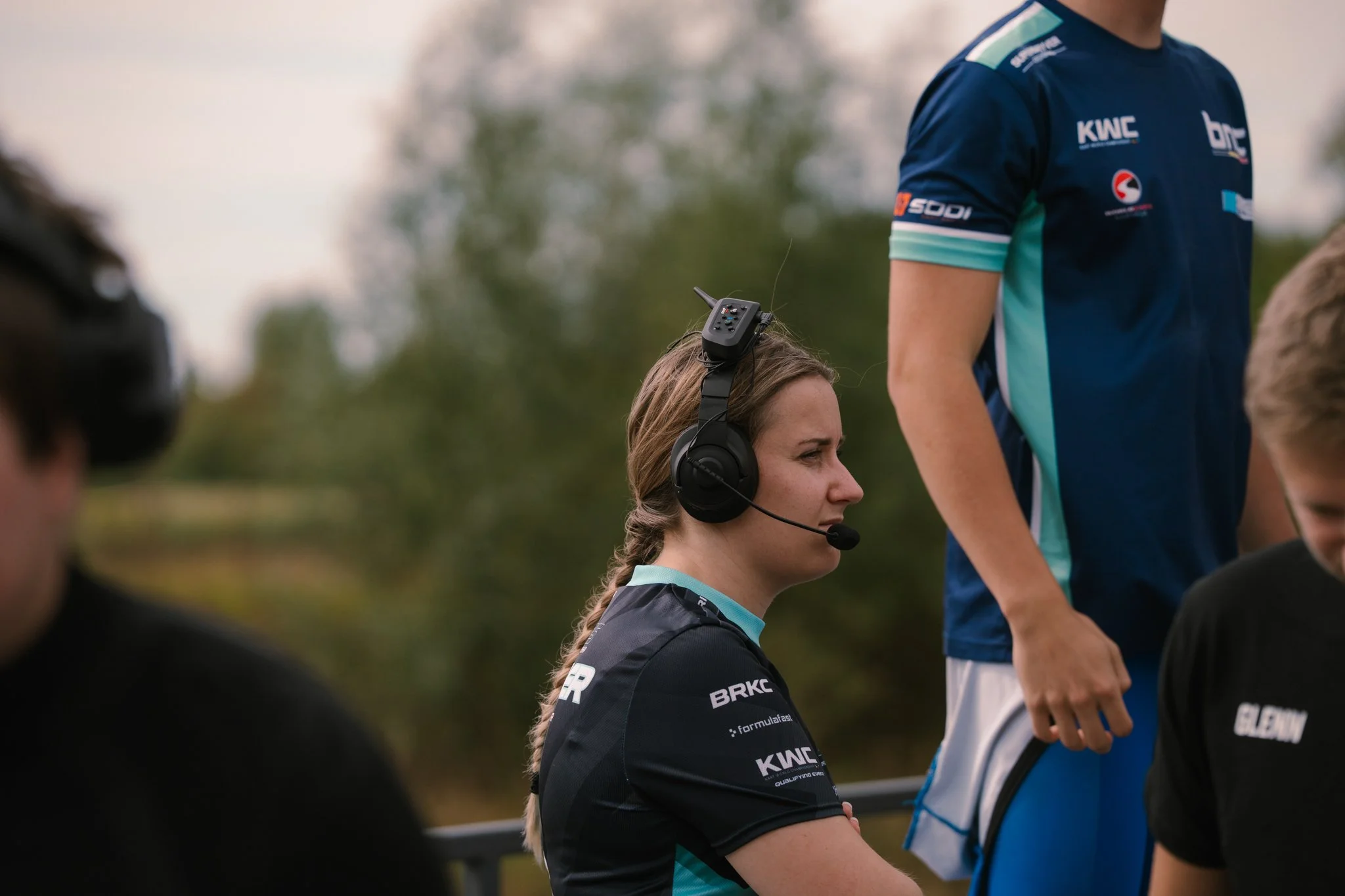 A woman wearing a black and teal sports shirt with sponsorship patches, a headset with a microphone, and braided hair, sitting outdoors with a serious expression. Partially visible are two young men standing nearby, one wearing a blue sports shirt an