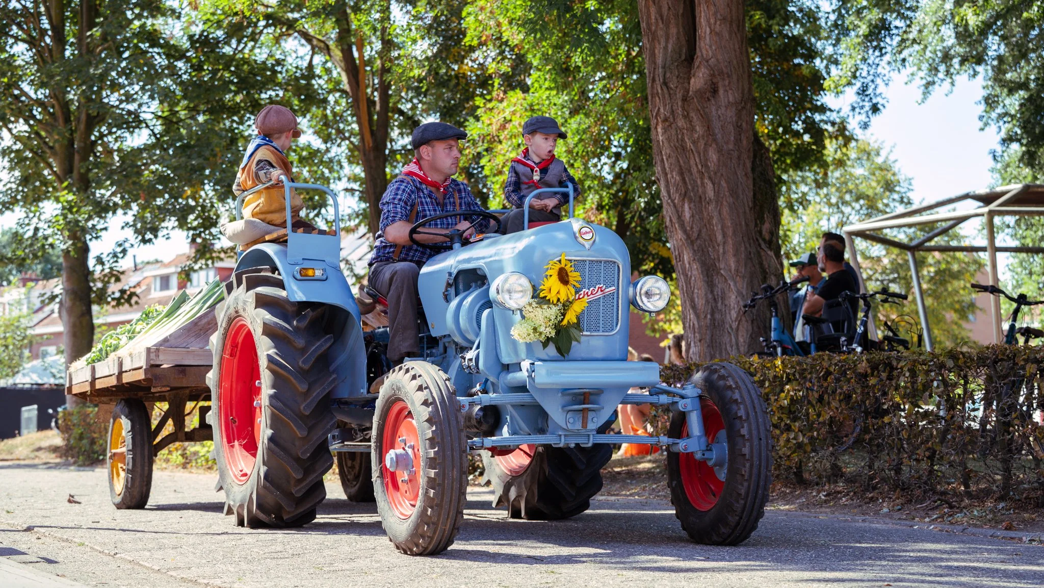 A vintage blue tractor decorated with sunflower and white flowers pulls a flatbed trailer carrying three children dressed as farmers with hats and boots, on a city street lined with trees and buildings.
