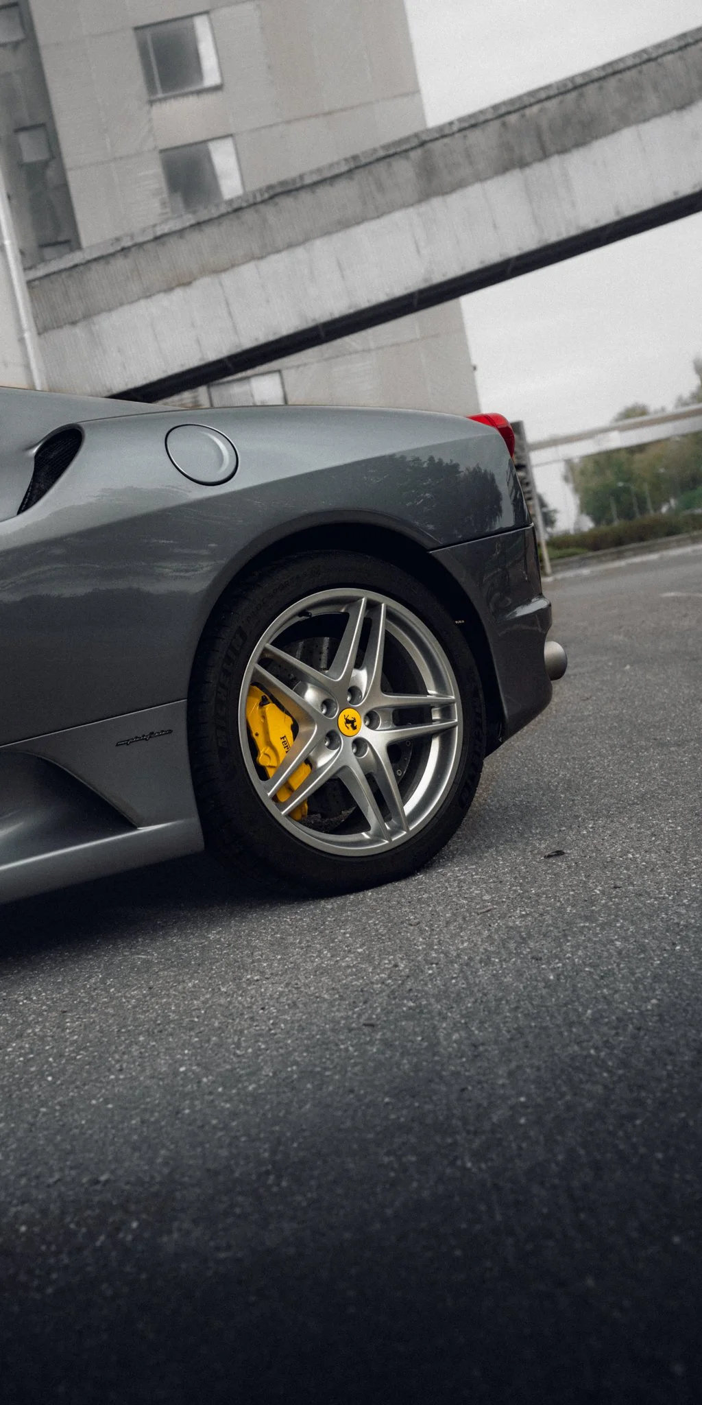 Close-up of a silver Ferrari car's front wheel, showing a yellow brake caliper with the Ferrari logo, parked on a gravel surface with buildings and overcast sky in the background.