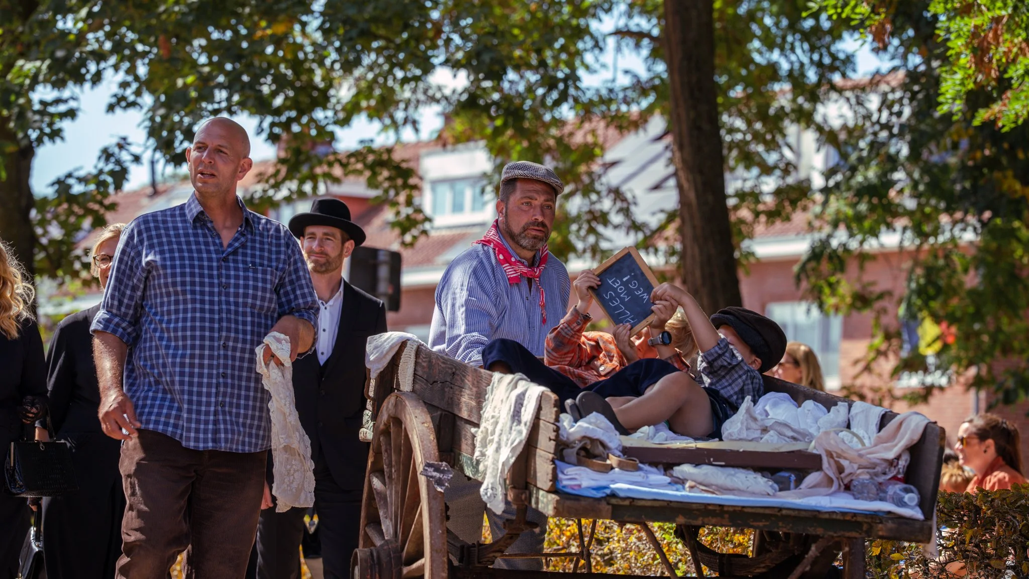 People in historical attire participating in a reenactment or parade outdoors, with some pulling a cart filled with linens and props, surrounded by trees and buildings.