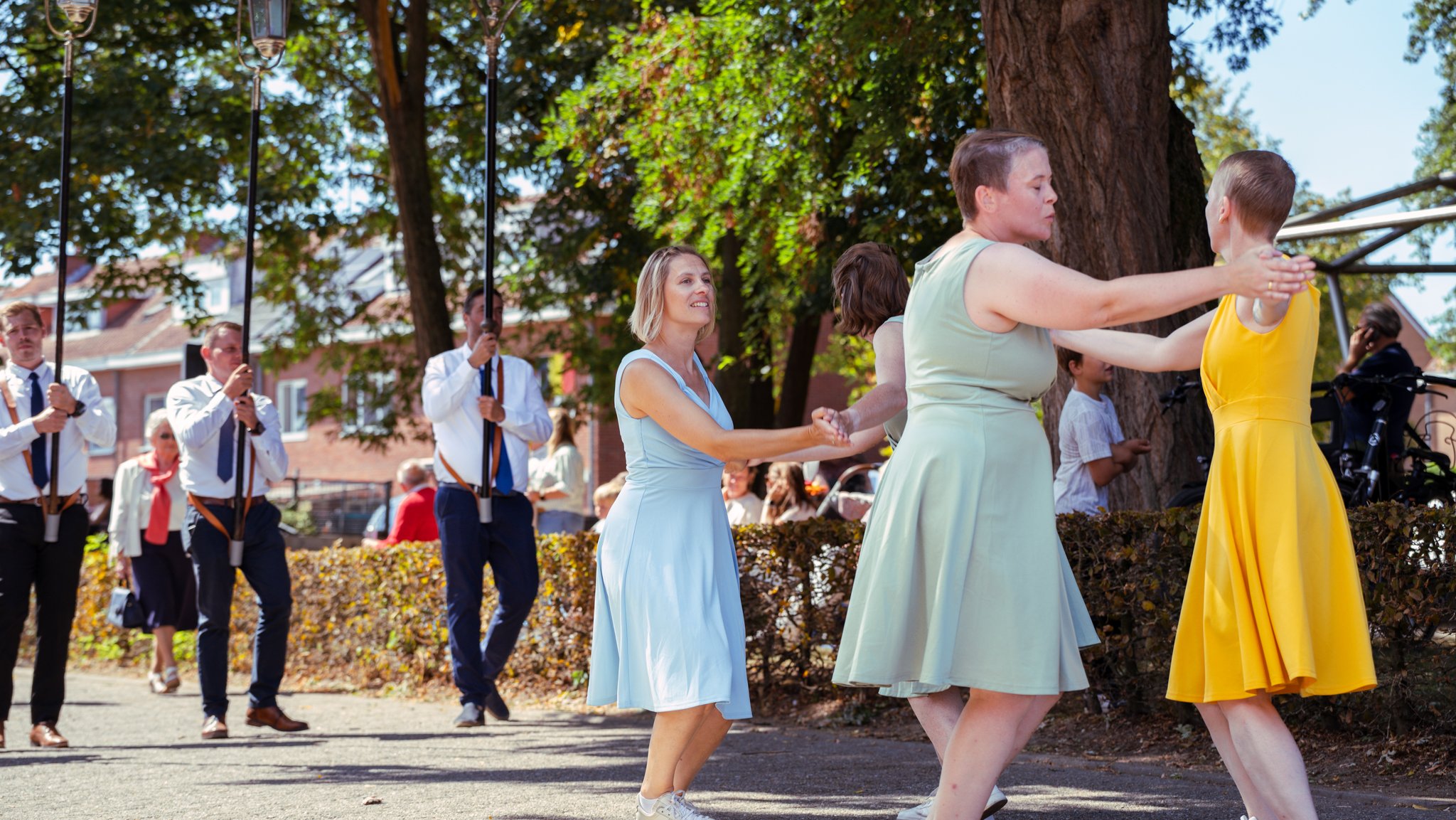 People dancing in a circle outdoors during a festival, with men holding flags in the background, and trees and buildings surrounding them.