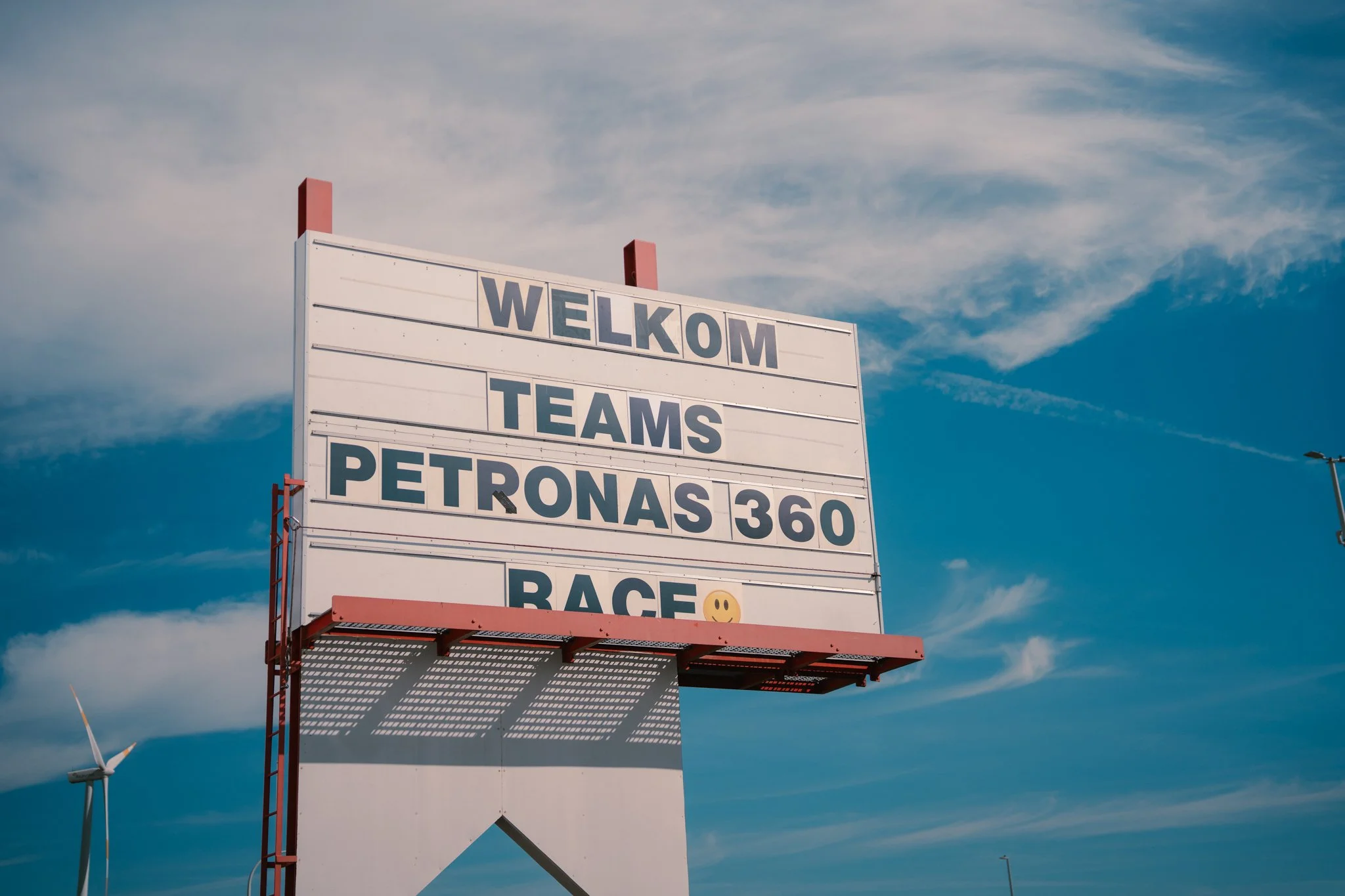 A large outdoor sign with a white background and black letters displaying the words: 'WELCOME TEAMS PETRONAS 360 RACE 😊', against a blue sky with scattered clouds.