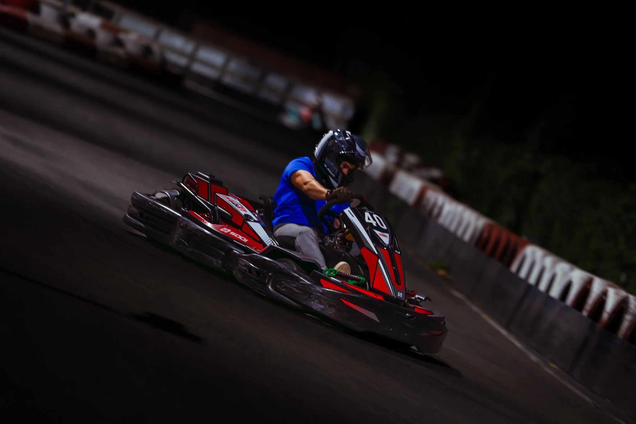 A person in a blue shirt and helmet driving a red and black go-kart on a race track at night.