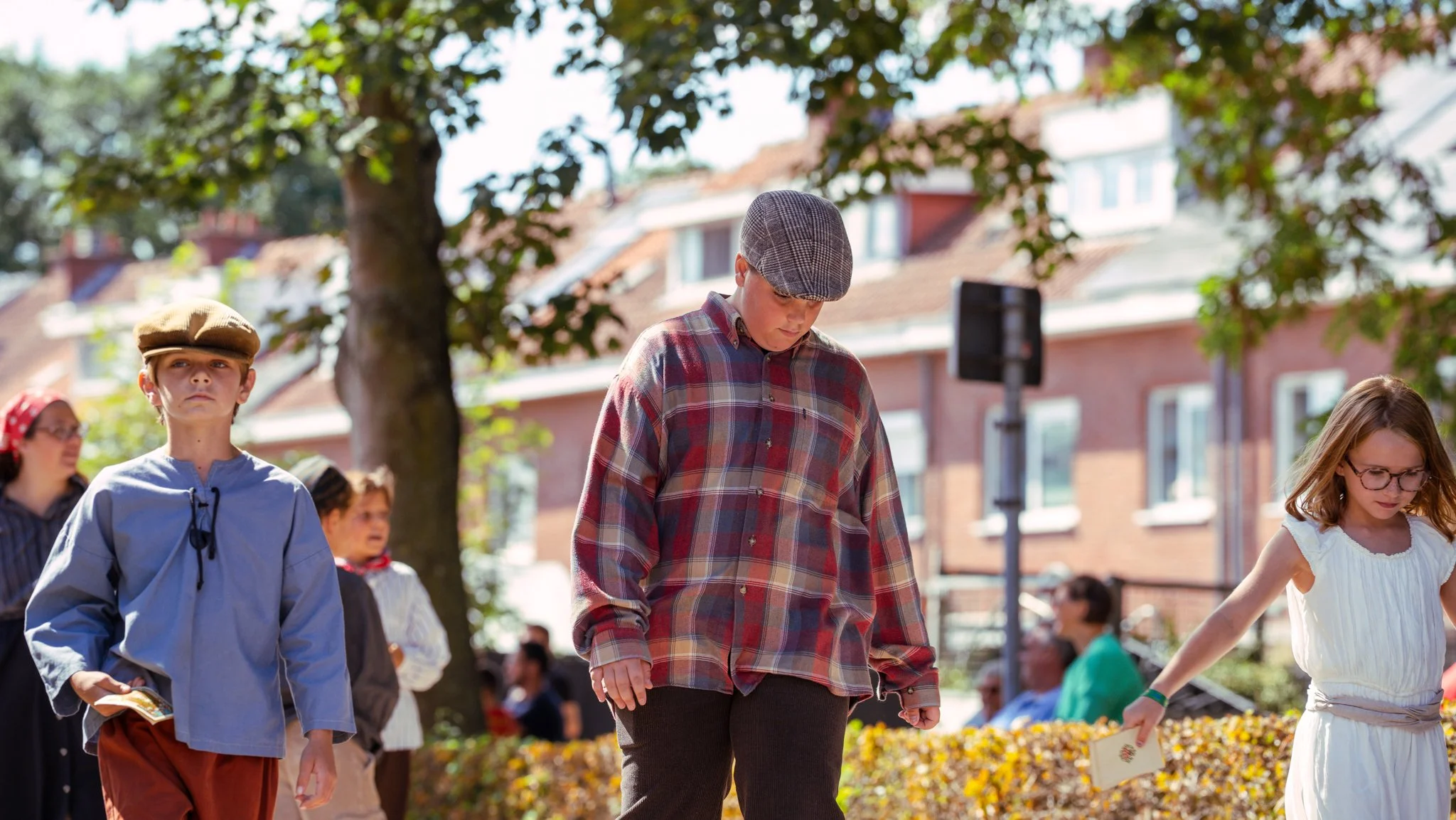 Children dressed in vintage clothing participating in an outdoor event on a sunny day, with trees and brick buildings in the background.