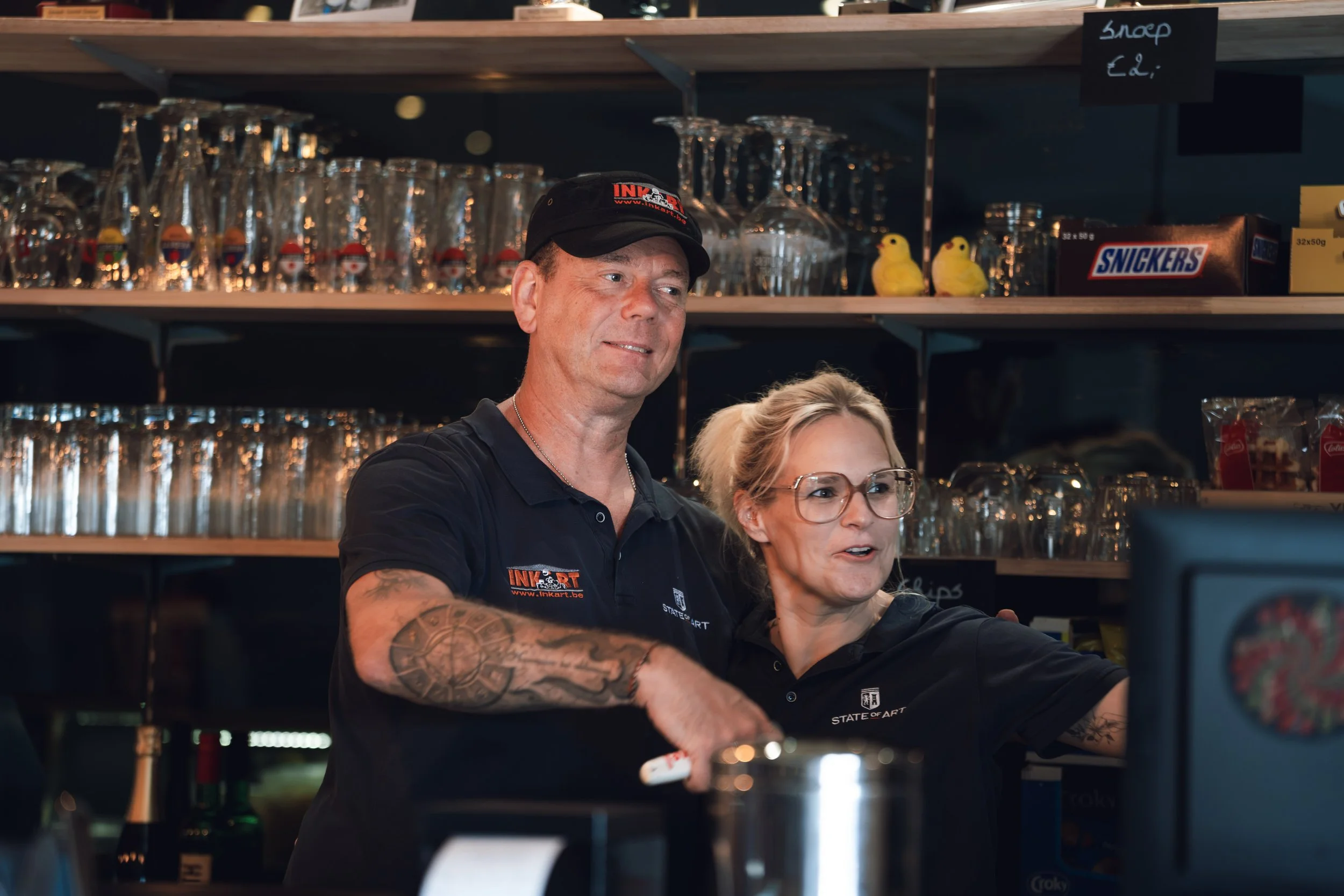 A man and a woman working together behind a bar counter, with shelves of glasses and a box of Snickers behind them.