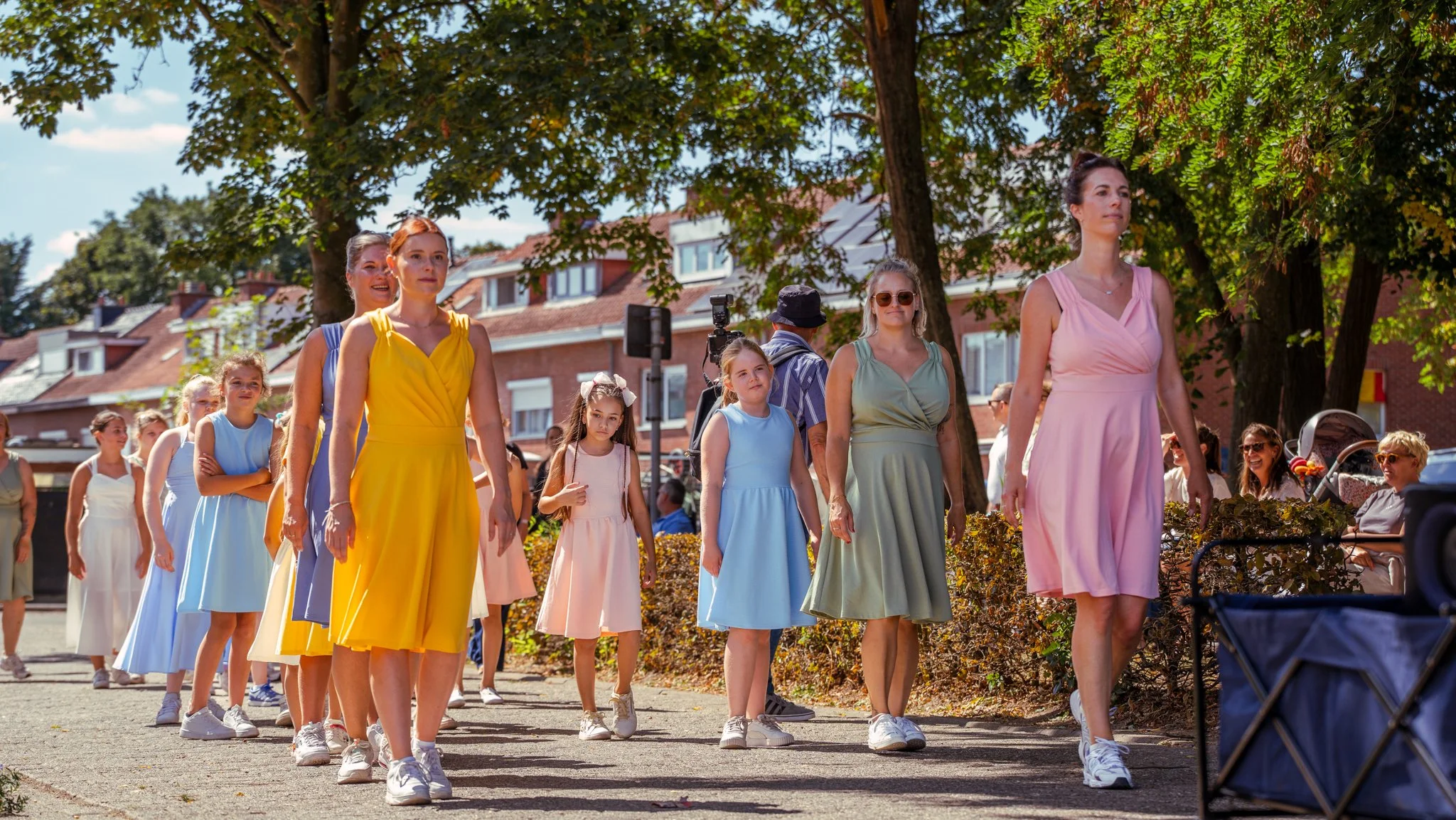 A group of young girls and women dressed in pastel-colored dresses are walking in a line outdoors on a sunny day, under a large tree, with houses in the background.