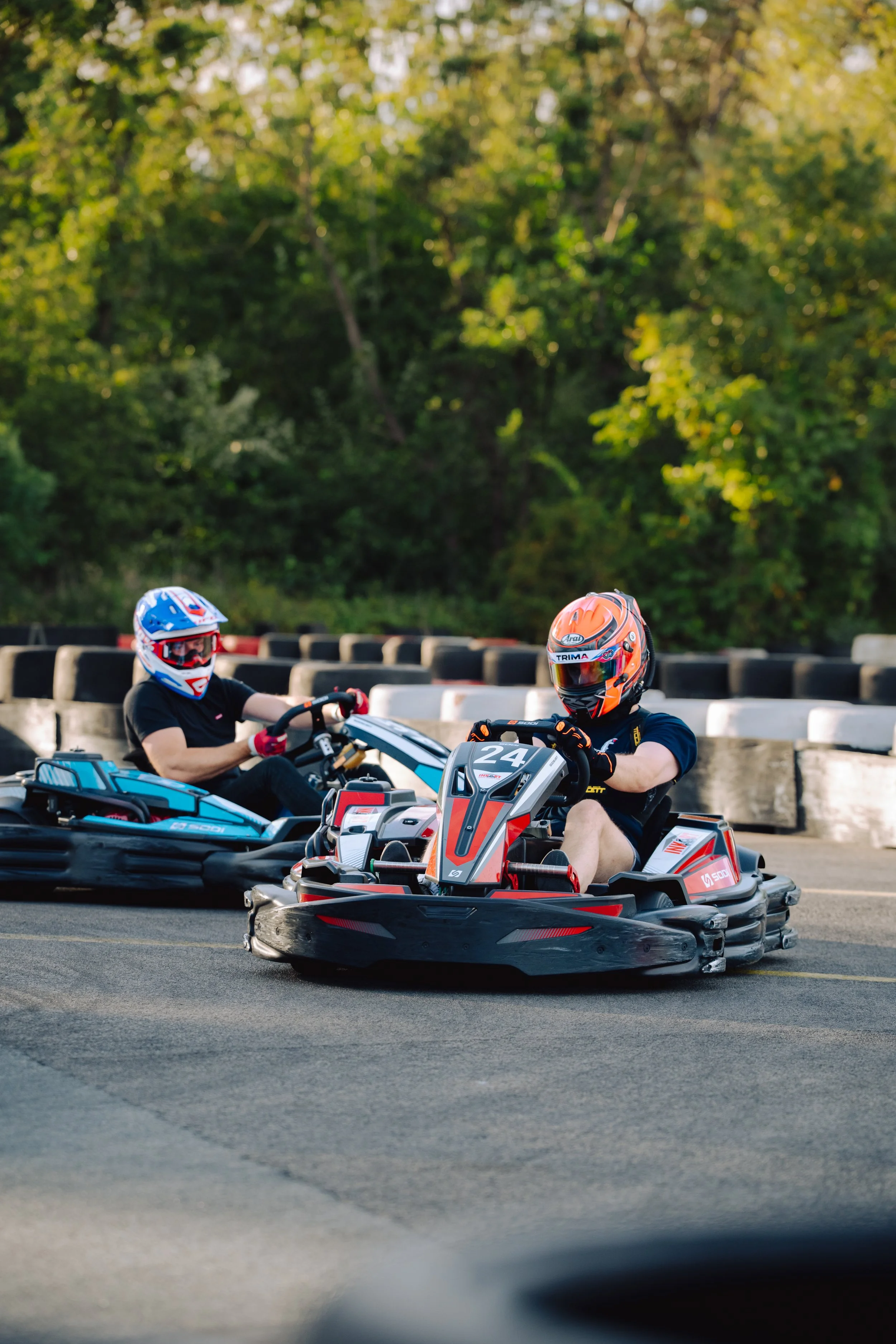 Two people riding go-karts on a track, wearing helmets and safety gear, with a background of trees and a barrier.