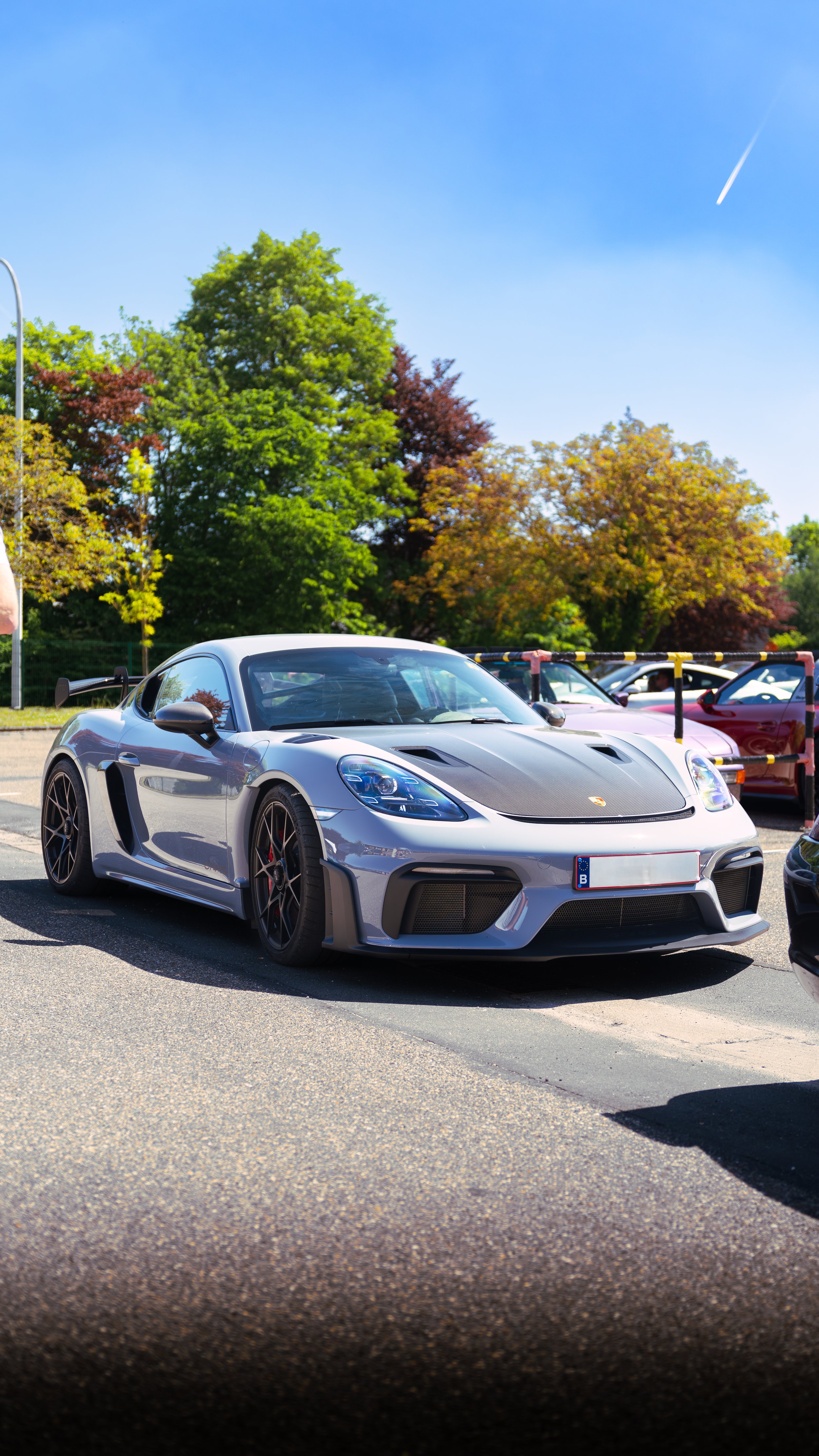 A gray sports car parked in a parking lot next to other vehicles, with green and fall-colored trees in the background, under a clear blue sky with a contrail.