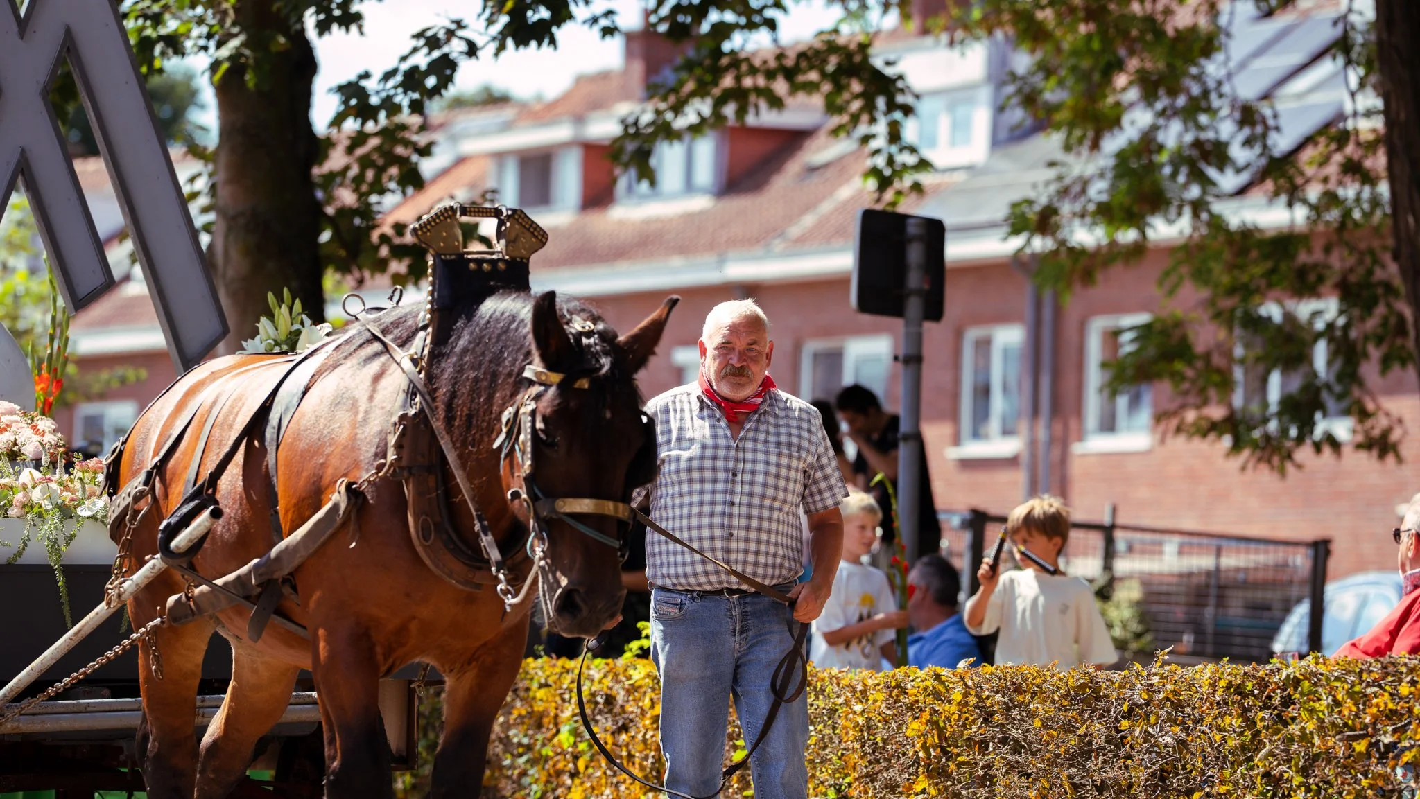 Man holding a horse's reins during a daytime outdoor event with children and a brick building in the background.