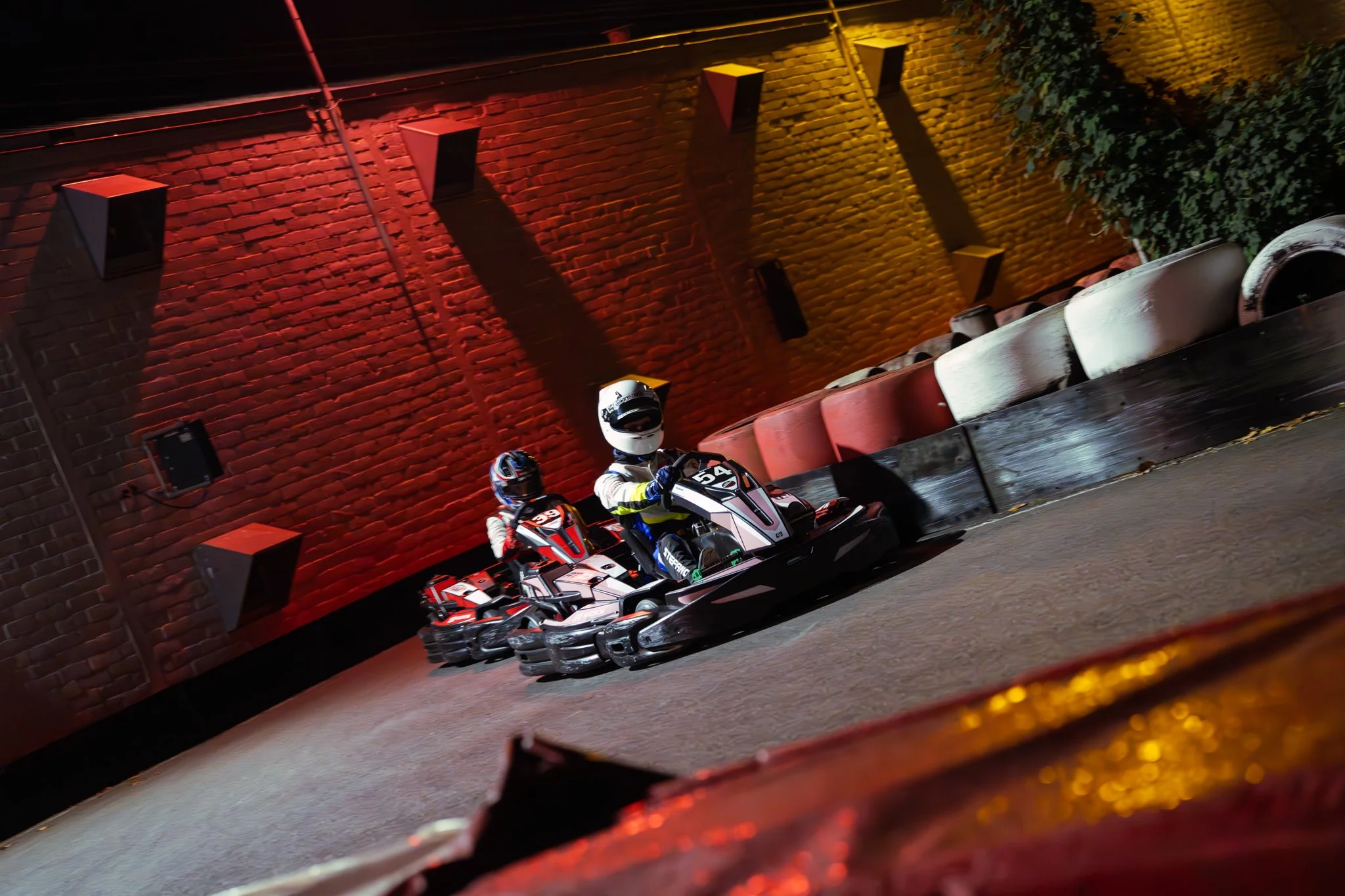 Two go-kart racers driving on an indoor track with a brick wall background, colorful tire barriers, and dramatic lighting.