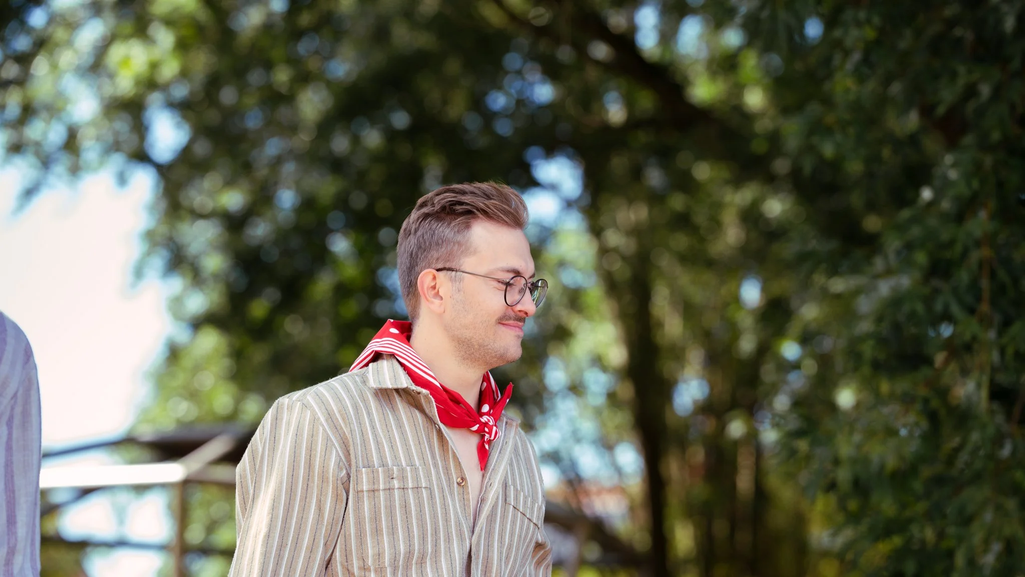 A young man with glasses and a red bandana around his neck, smiling with his eyes closed outdoors surrounded by trees.