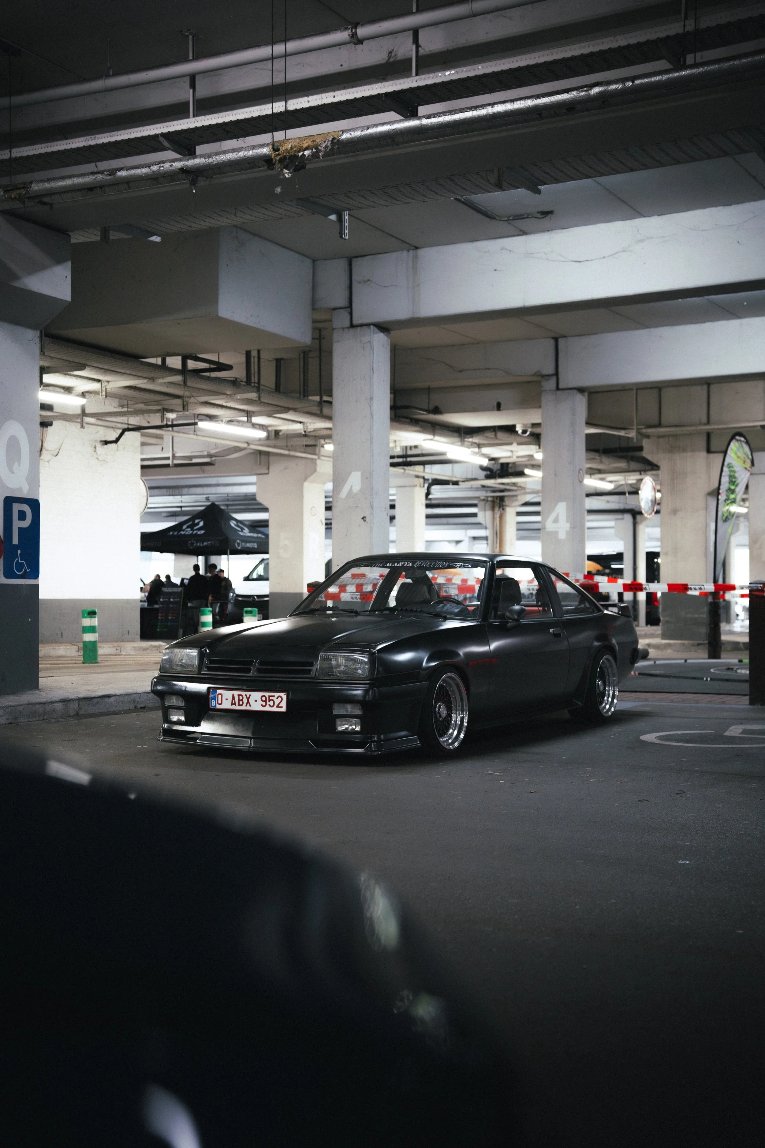 A black vintage sports car parked in an underground parking garage with concrete pillars and ceiling pipes. There are some people in the background near a black tent and parking signs.